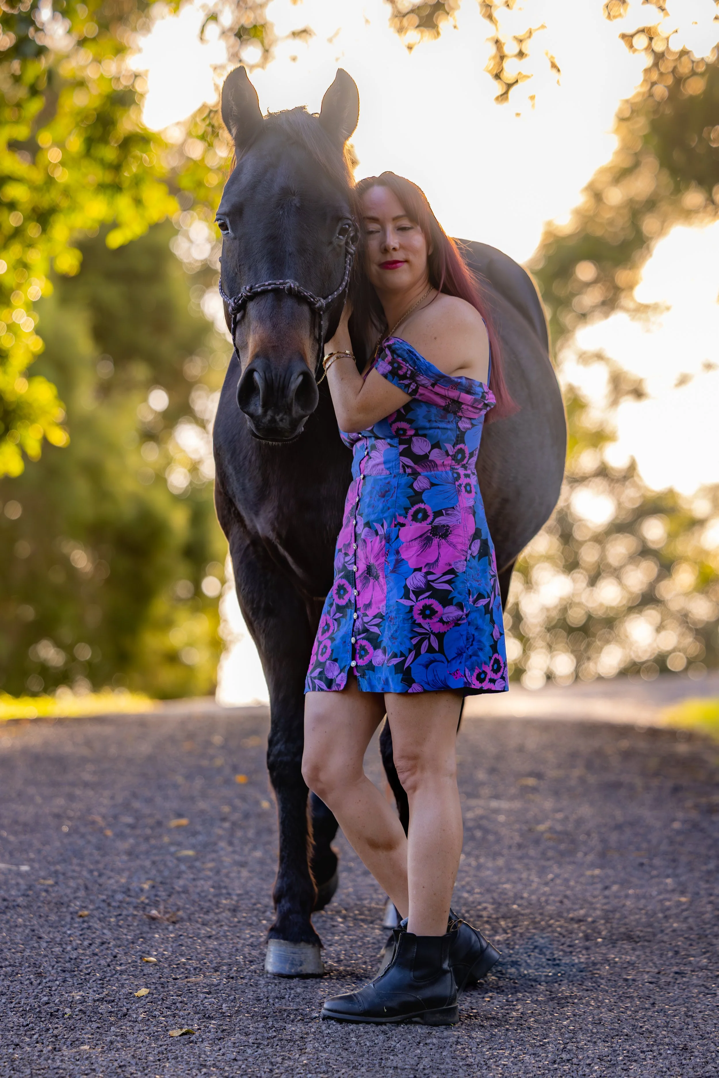 A woman with long dark hair wearing a blue and purple floral dress and black combat boots standing close to a black horse with a halter in a sunlit outdoor area with trees and a gravel path.
