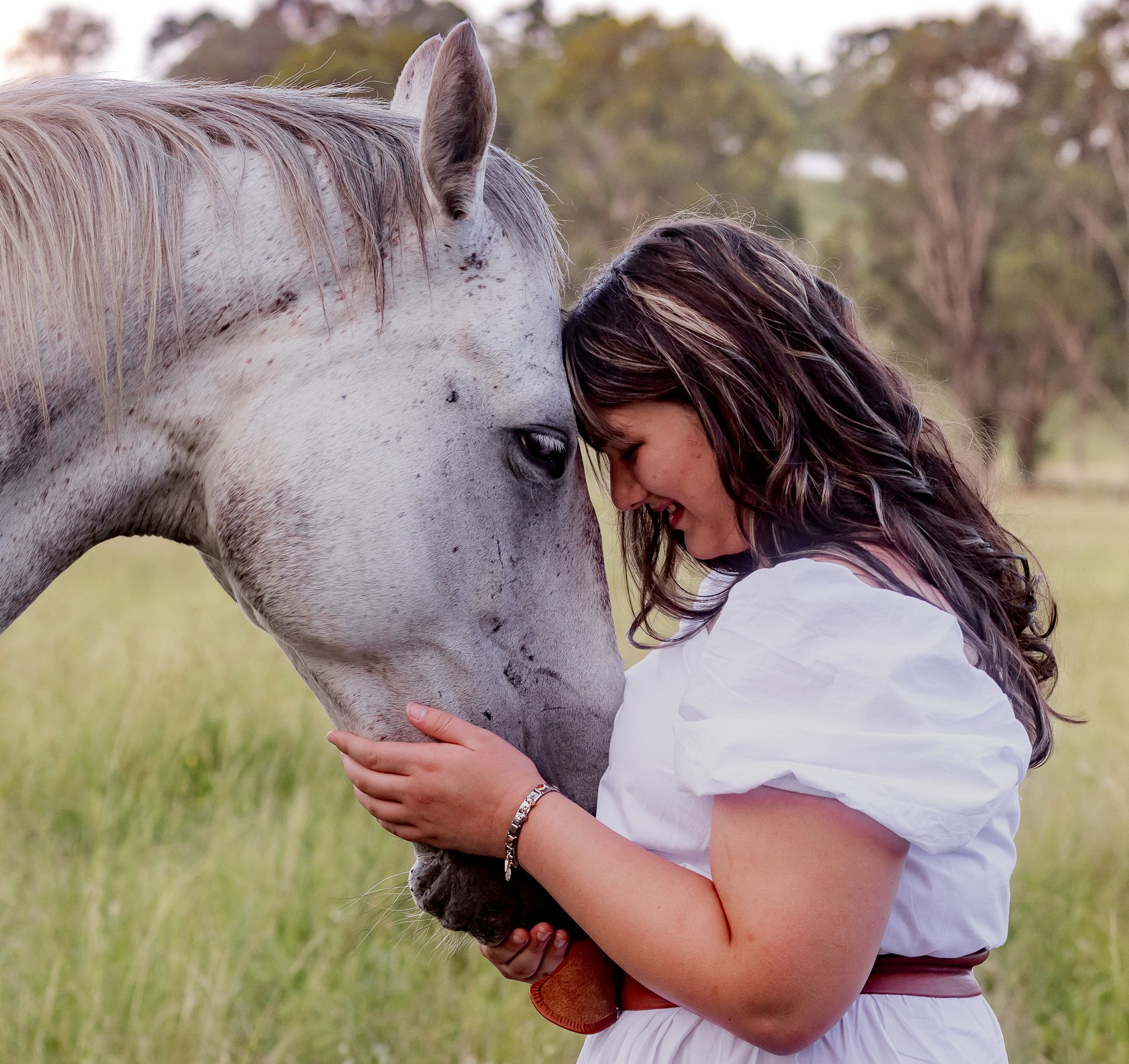 A woman gently touching foreheads and smiling with a white horse in a grassy field.
