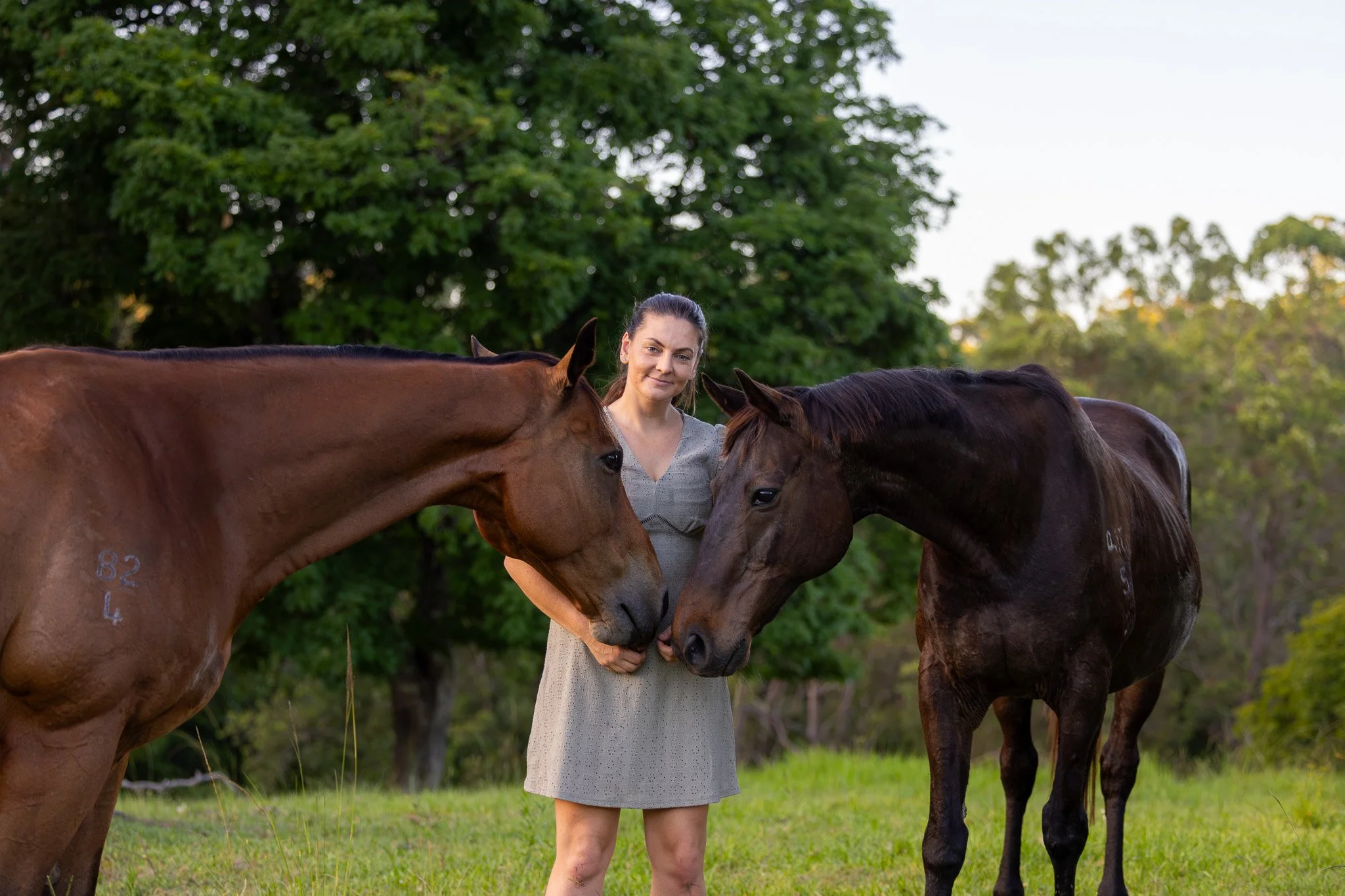 A woman standing outdoors in a grassy area, holding two horses by their heads, one chestnut and one dark brown, with green trees in the background.