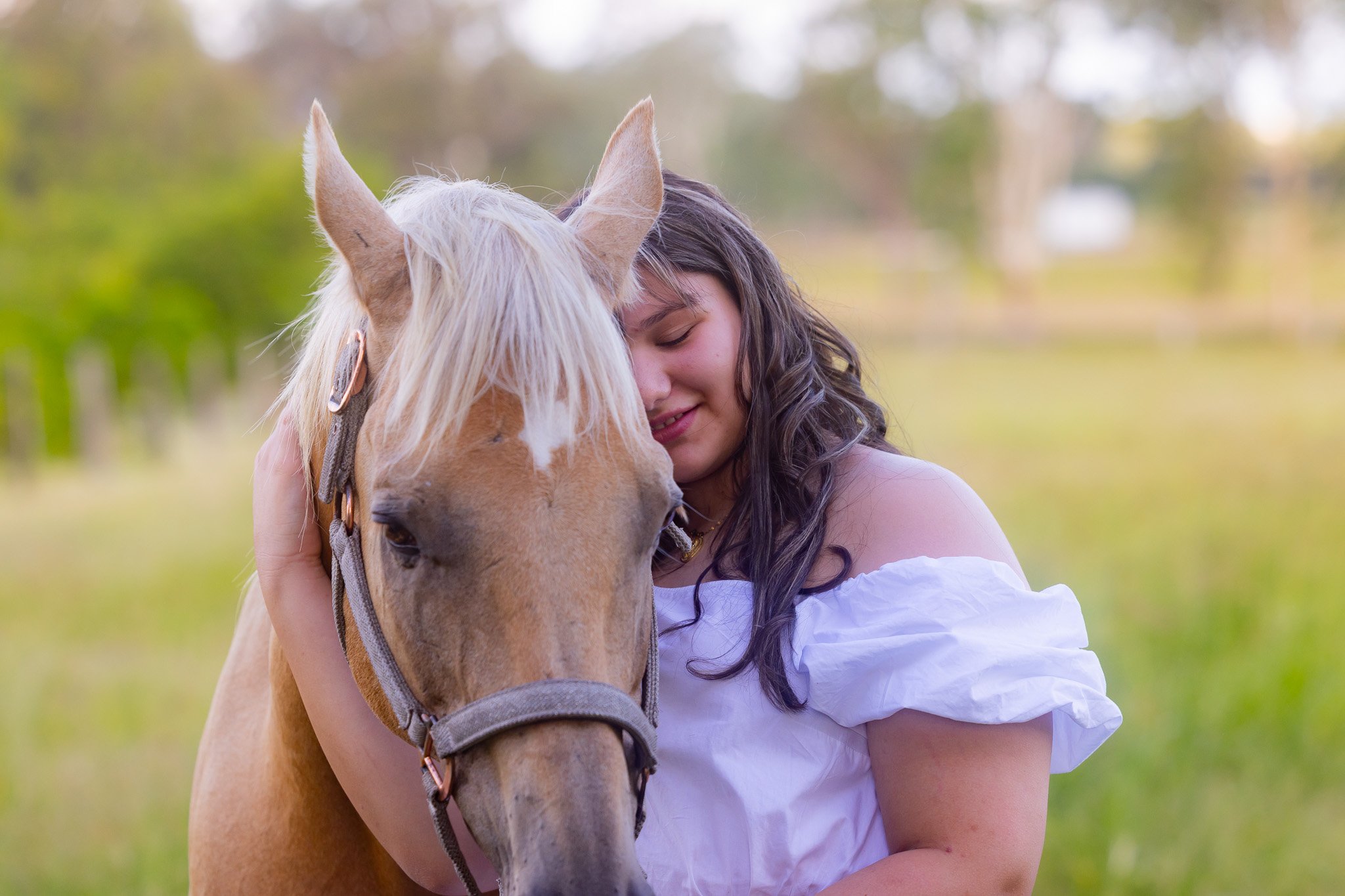 Woman in white dress hugging a tan horse with a bridle in a grassy field.