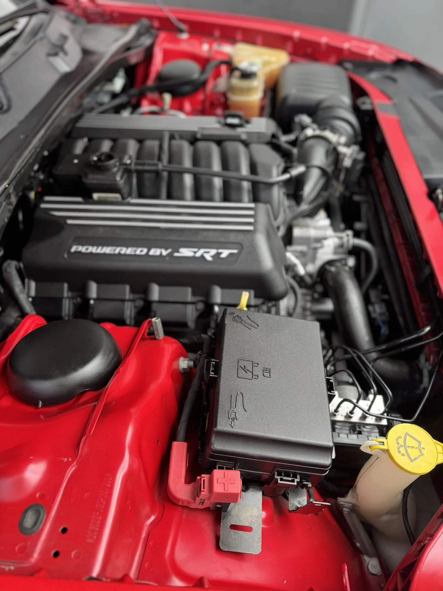 Close-up of a car engine compartment with "Powered by SRT" branding on the engine cover, surrounded by red chassis components and various engine parts, including a coolant reservoir with a yellow cap.