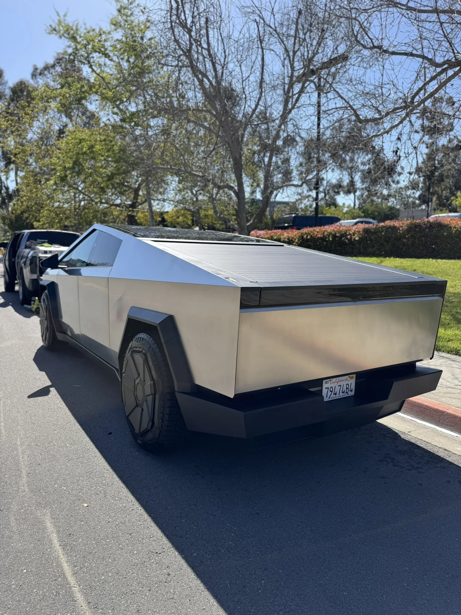 A futuristic, angular electric vehicle parked on a street, surrounded by trees and grassy area under a clear blue sky.