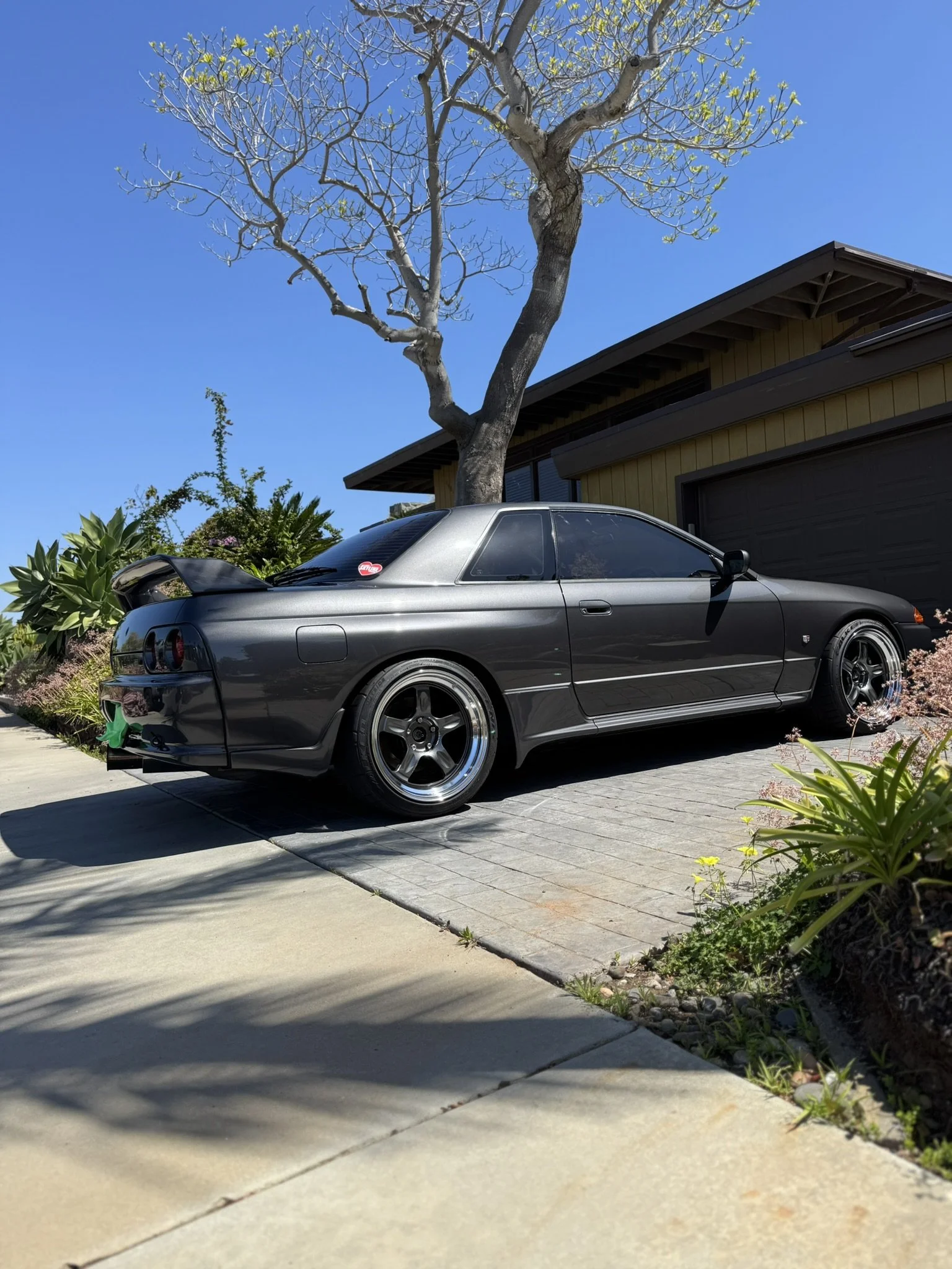 Gray sports car parked on a driveway beside a house with a large tree and clear blue sky in the background.