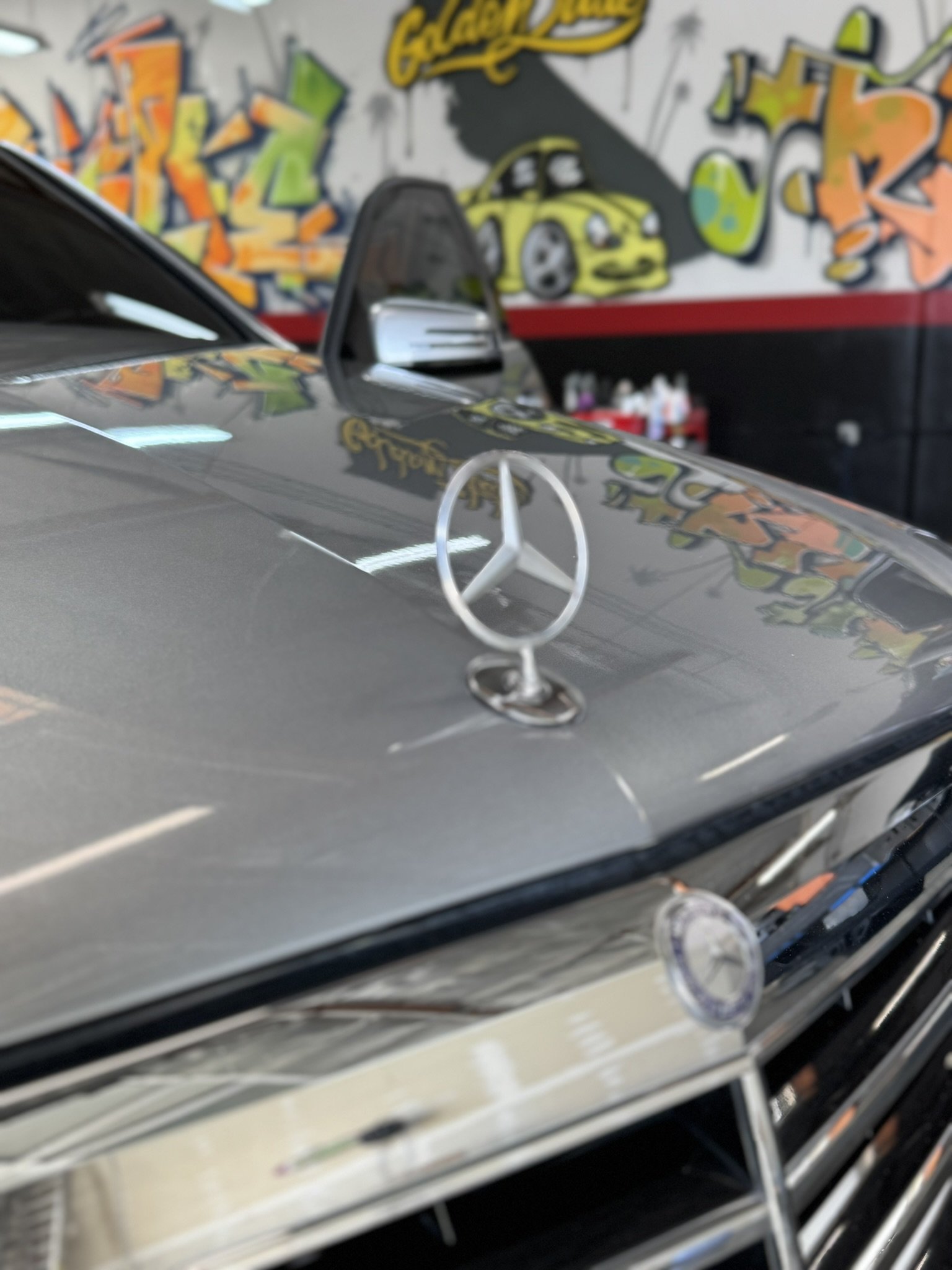 Close-up of a Mercedes-Benz hood ornament with a graffiti-covered wall in the background.