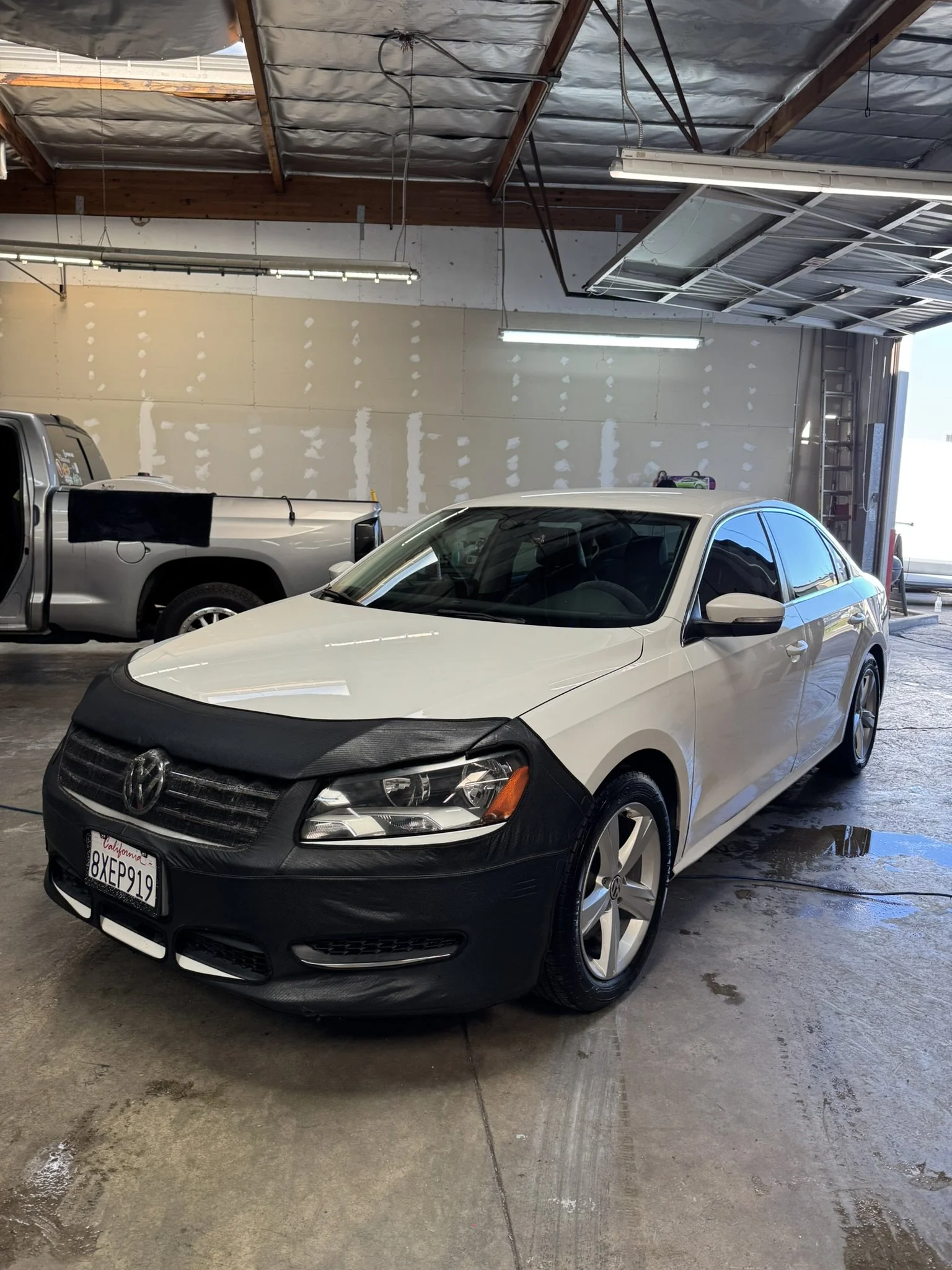 A white Volkswagen car with a black front bra cover inside a garage, next to a silver pickup truck.