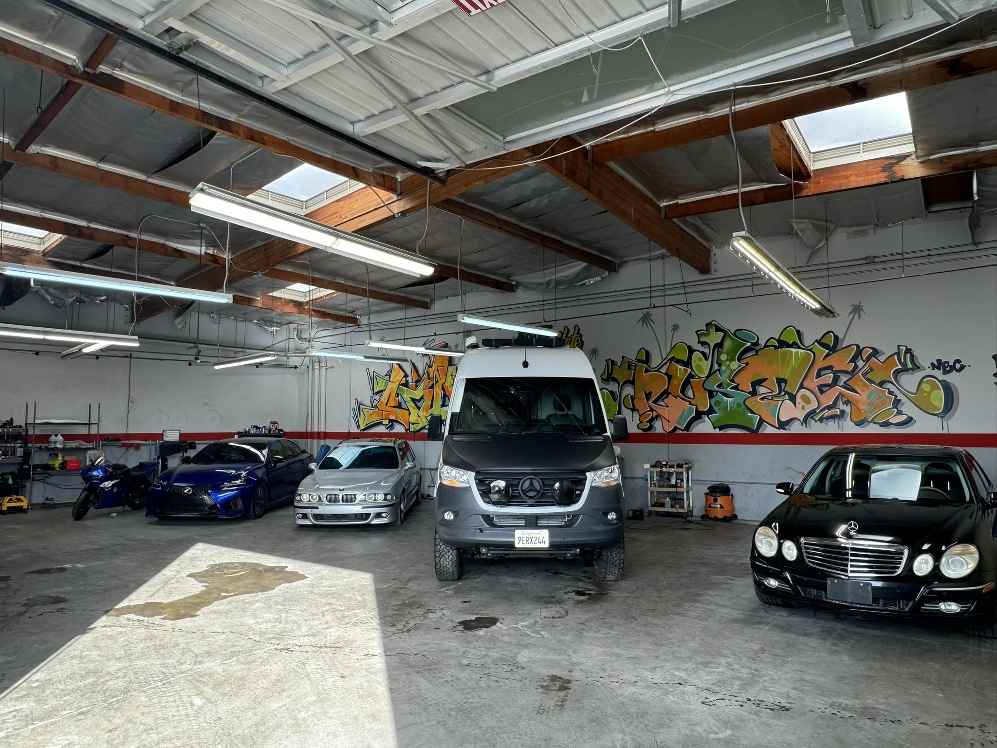 Interior of a garage with multiple vehicles, including a white van, two cars, and a motorcycle. The walls have colorful graffiti art, and there are overhead lights and exposed beams.