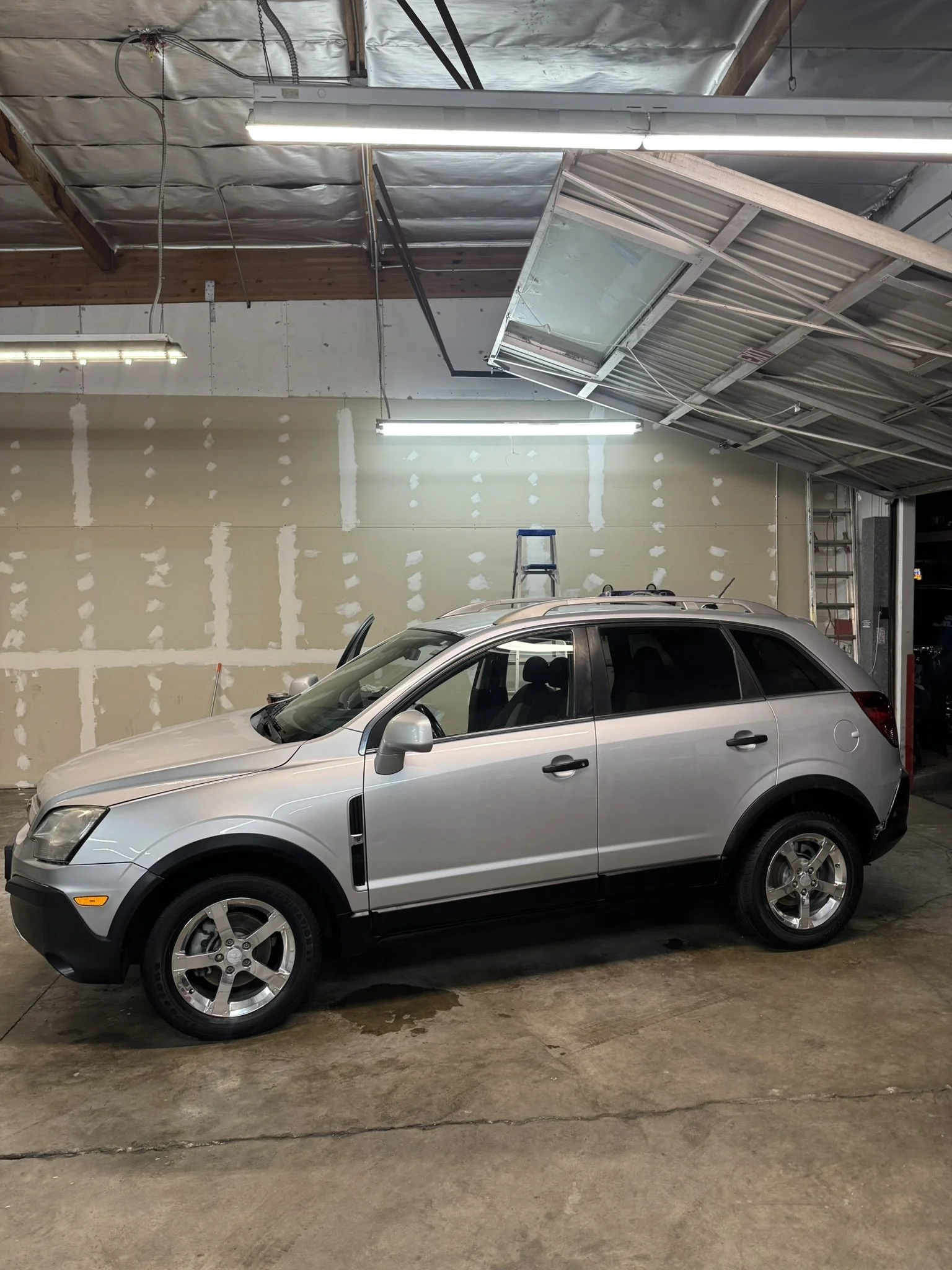 Silver SUV parked in a garage with partially open garage door, industrial lighting, and visible wall repairs.