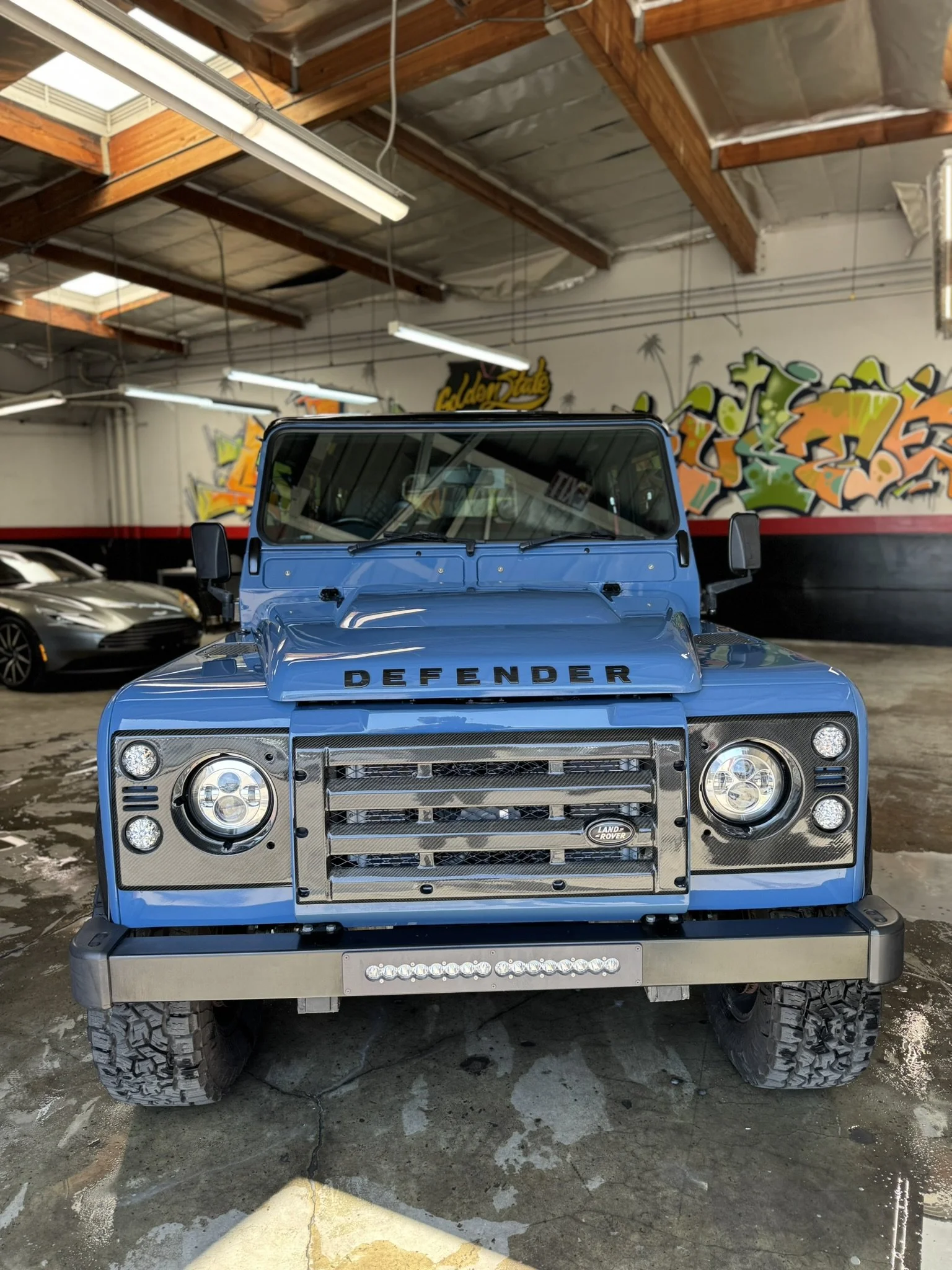 Blue Land Rover Defender parked in a garage with graffiti on the walls and wooden beams on the ceiling.