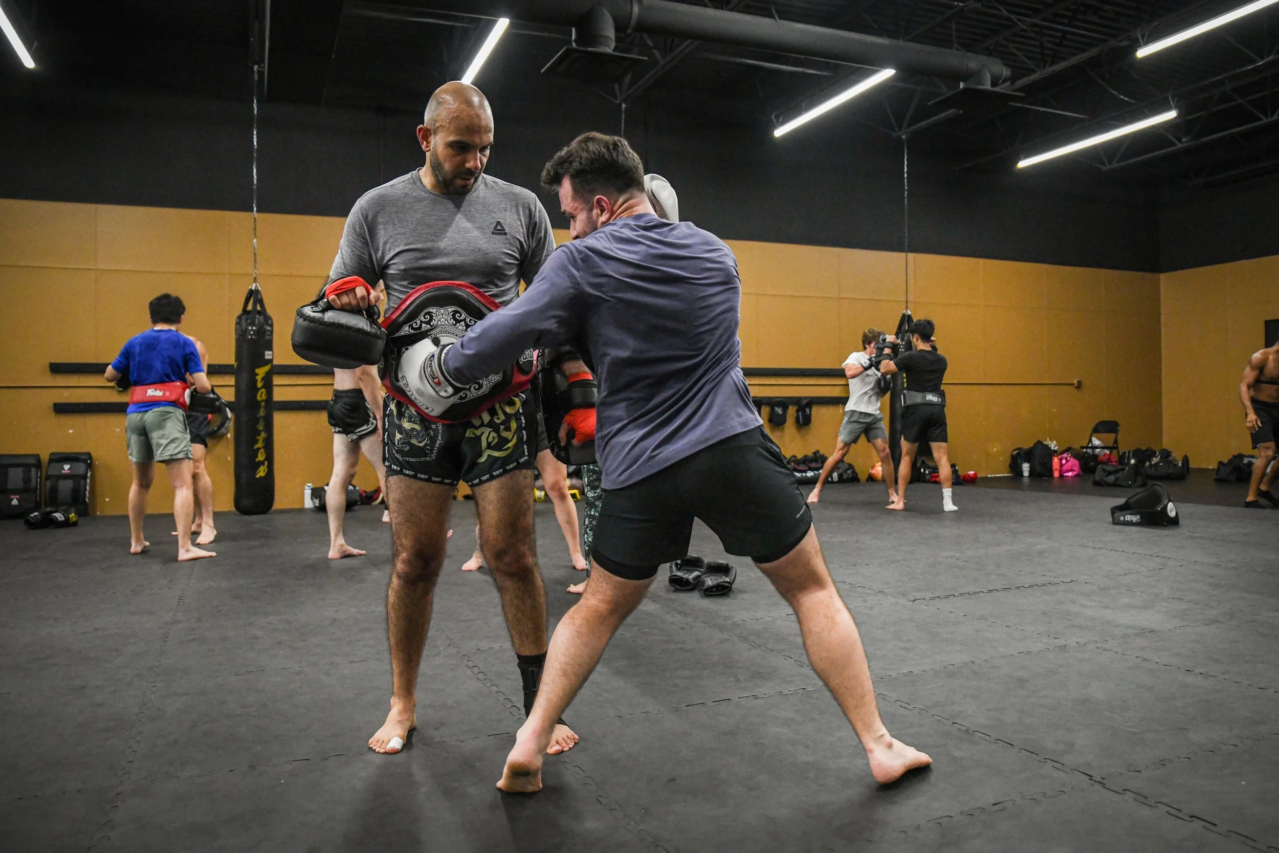 A large group of individuals practicing martial arts in a gym, using focus mitts and pads for training, with mirrors along one wall and punching bags hanging in the background.