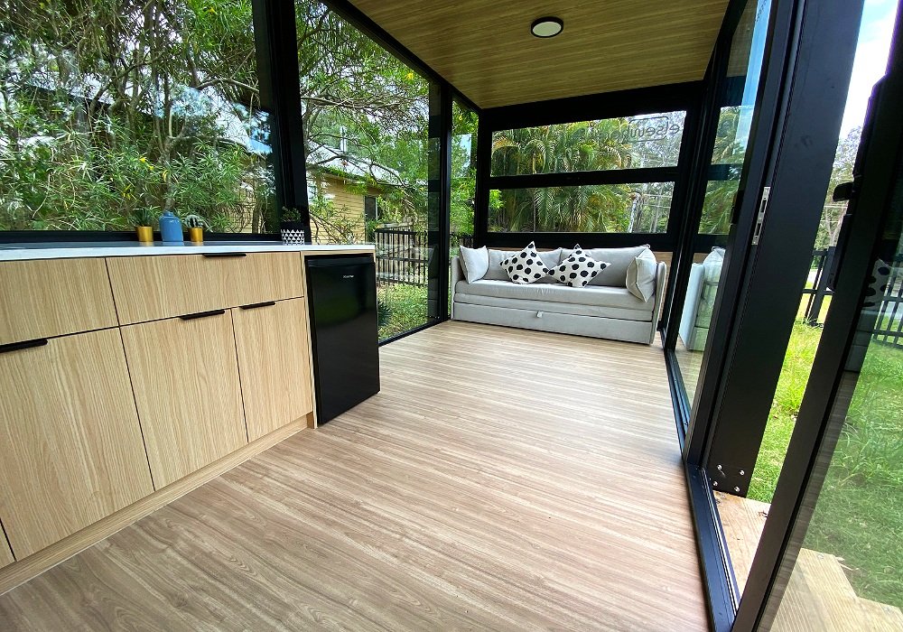Sleek and functional kitchen featuring custom cabinetry, stone benchtops, and a bar fridge. The open-plan design seamlessly connects the living space, creating a bright and airy atmosphere with floor-to-ceiling glass panels.