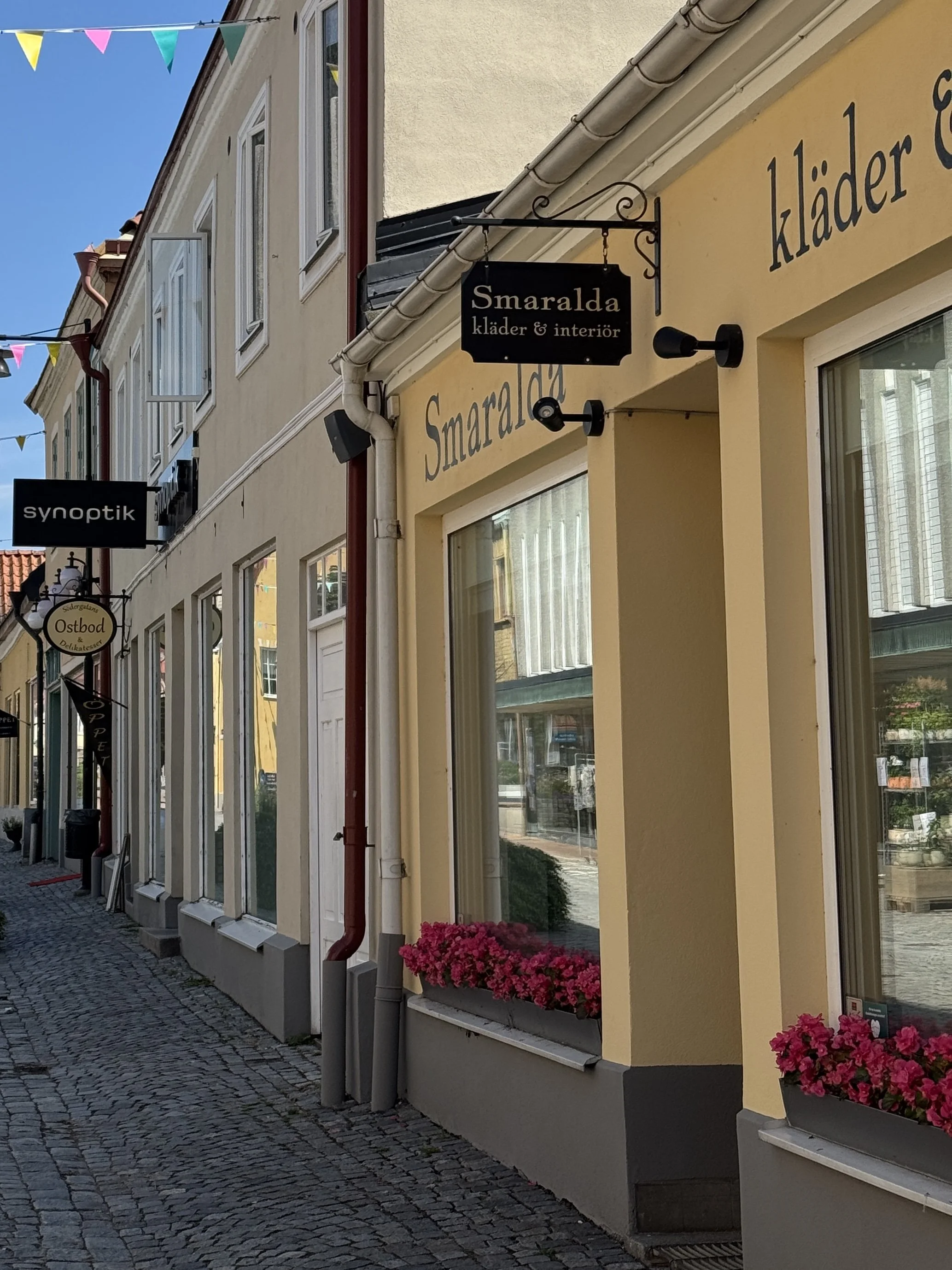 Skylt för butik 'Smaralda kläder & interiör' på en stadsgata, vy mot andra butiker och byggnader i bakgrunden.