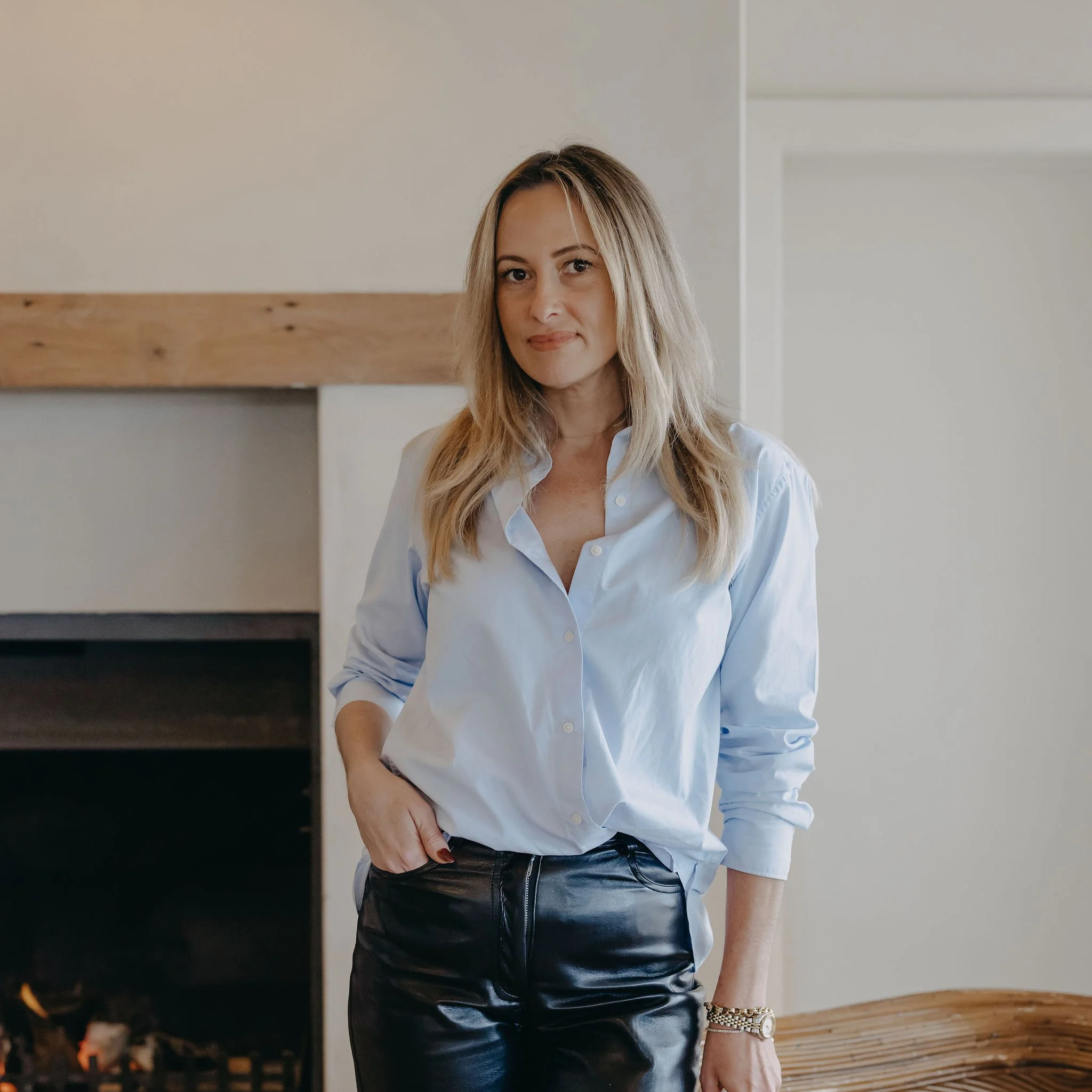 A woman with long blonde hair standing in front of a fireplace, wearing a light blue button-up shirt and black leather pants.