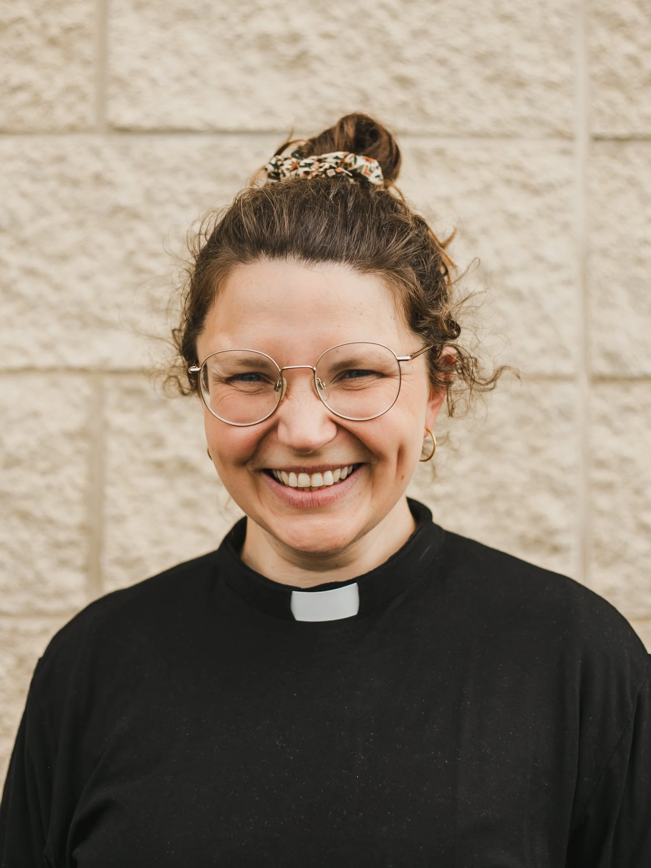 Imogen Ball, clergy at Orchard Grove Church. A smiling woman wearing glasses and a black clergy shirt with a clerical collar, standing against a beige brick wall.