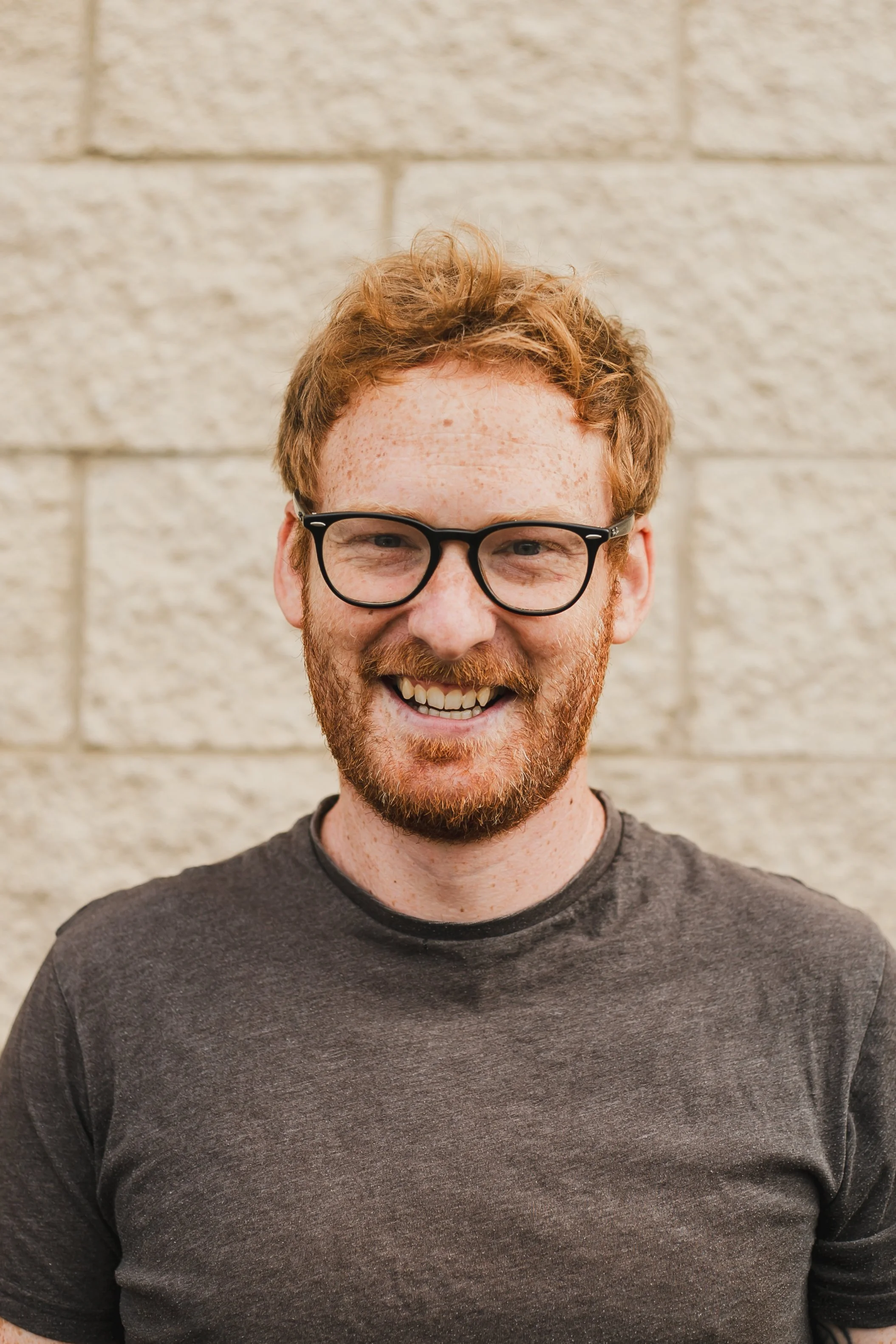 Lawrence Arnold. A smiling man with red hair, glasses, and a beard stands in front of a beige brick wall.