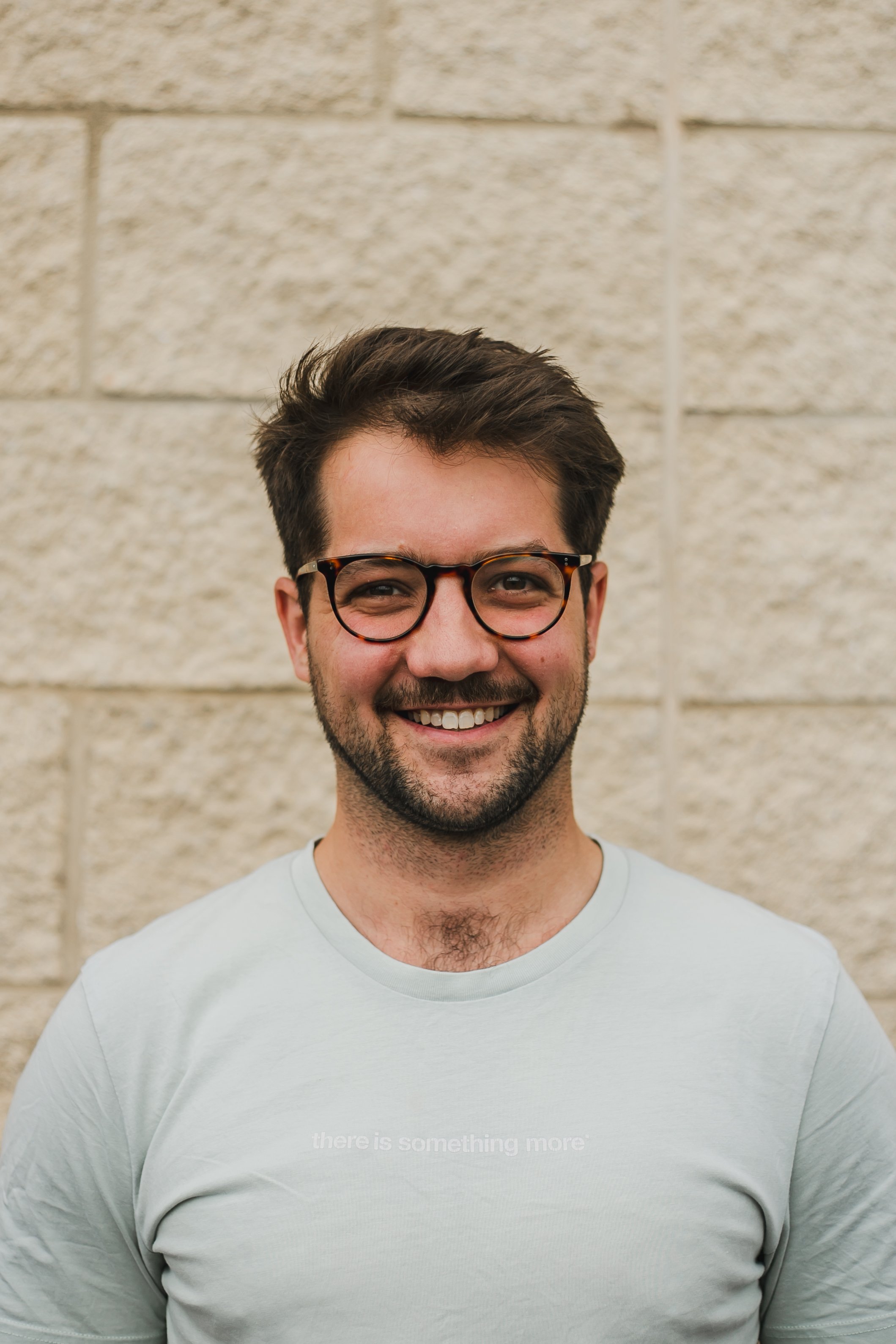 Matt Bodey. A smiling man with dark hair, wearing glasses and a light-colored t-shirt, standing in front of a beige brick wall.