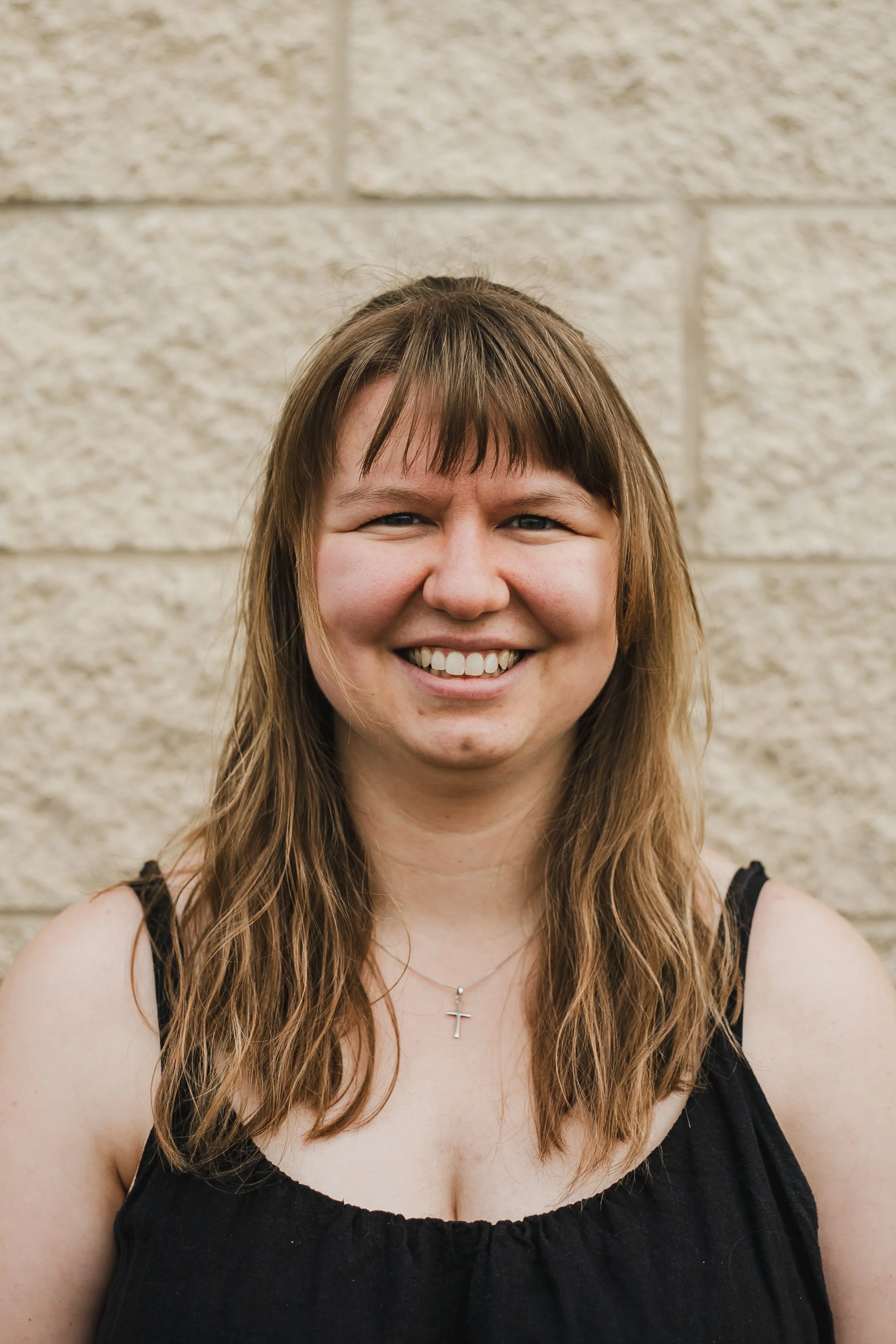 Megan Jones-Dellaportas. A smiling woman with shoulder-length light brown hair, wearing a black sleeveless top and a silver necklace with a cross pendant, standing in front of a beige brick wall.