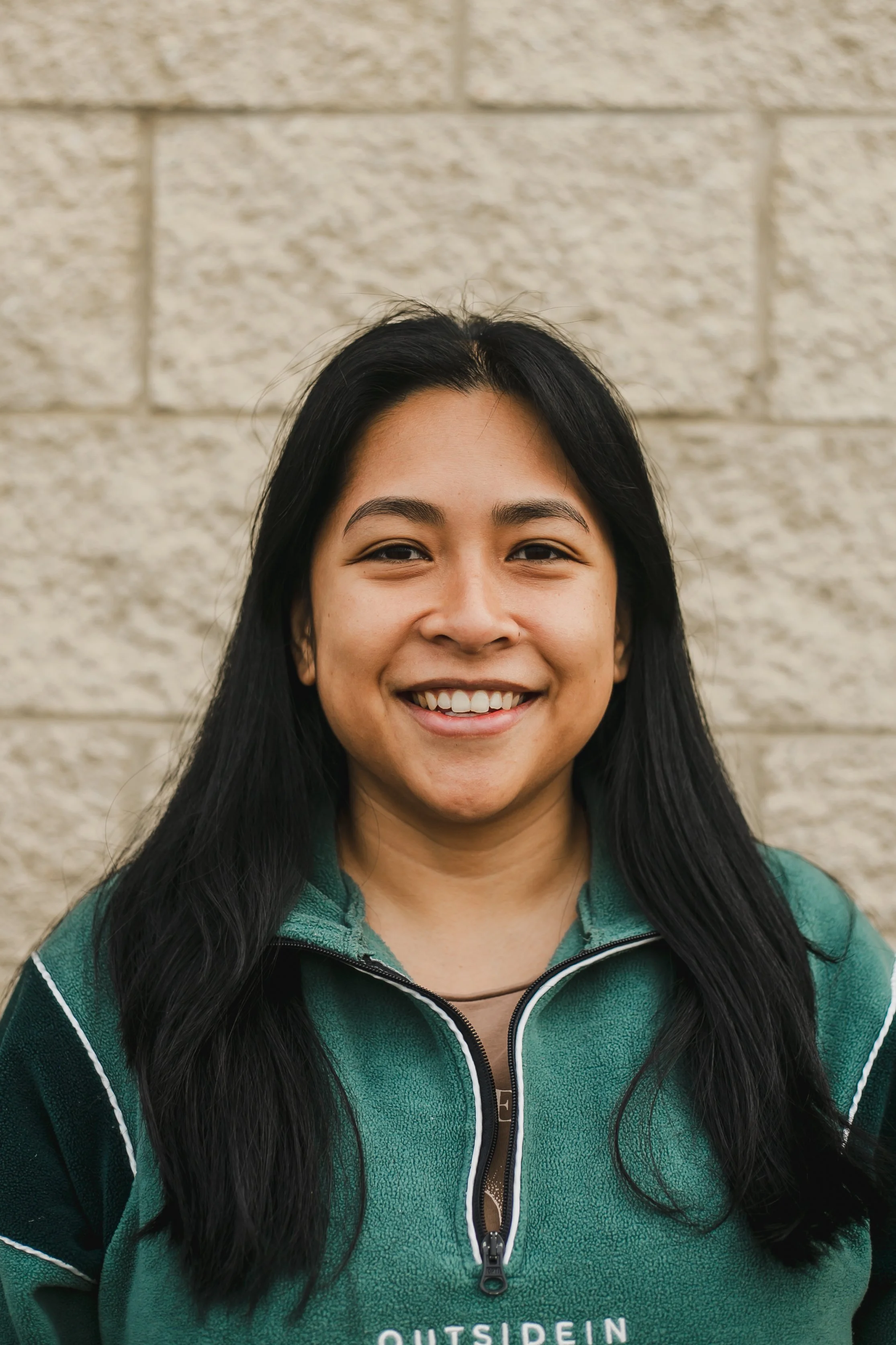Marjay Pallant. Young woman with long black hair smiling, standing against a beige brick wall, wearing a green zip-up jacket.