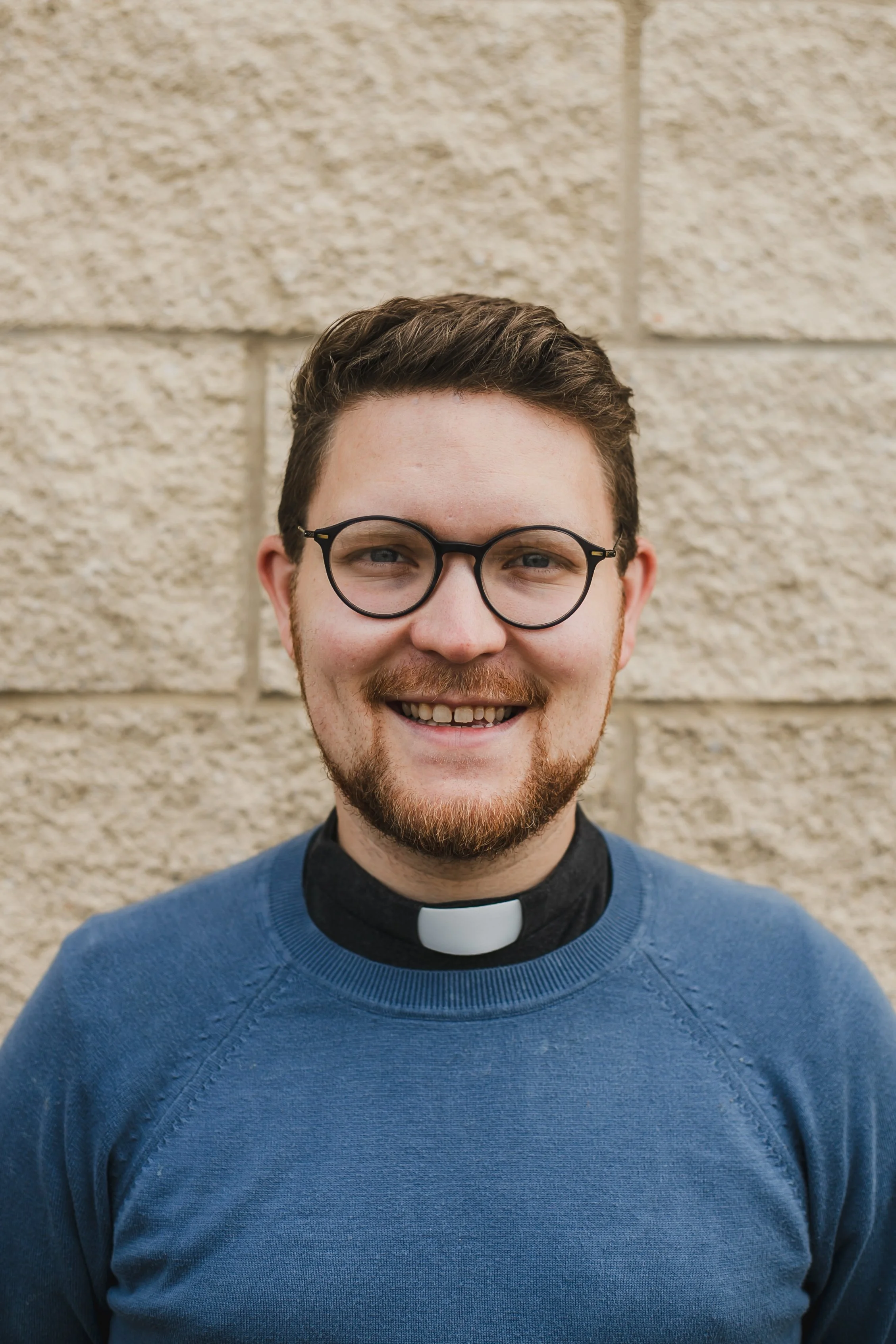 Jon Ball, vicar at Orchard Grove church. Smiling man wearing glasses, clergy collar, and a blue sweater standing against a beige brick wall.