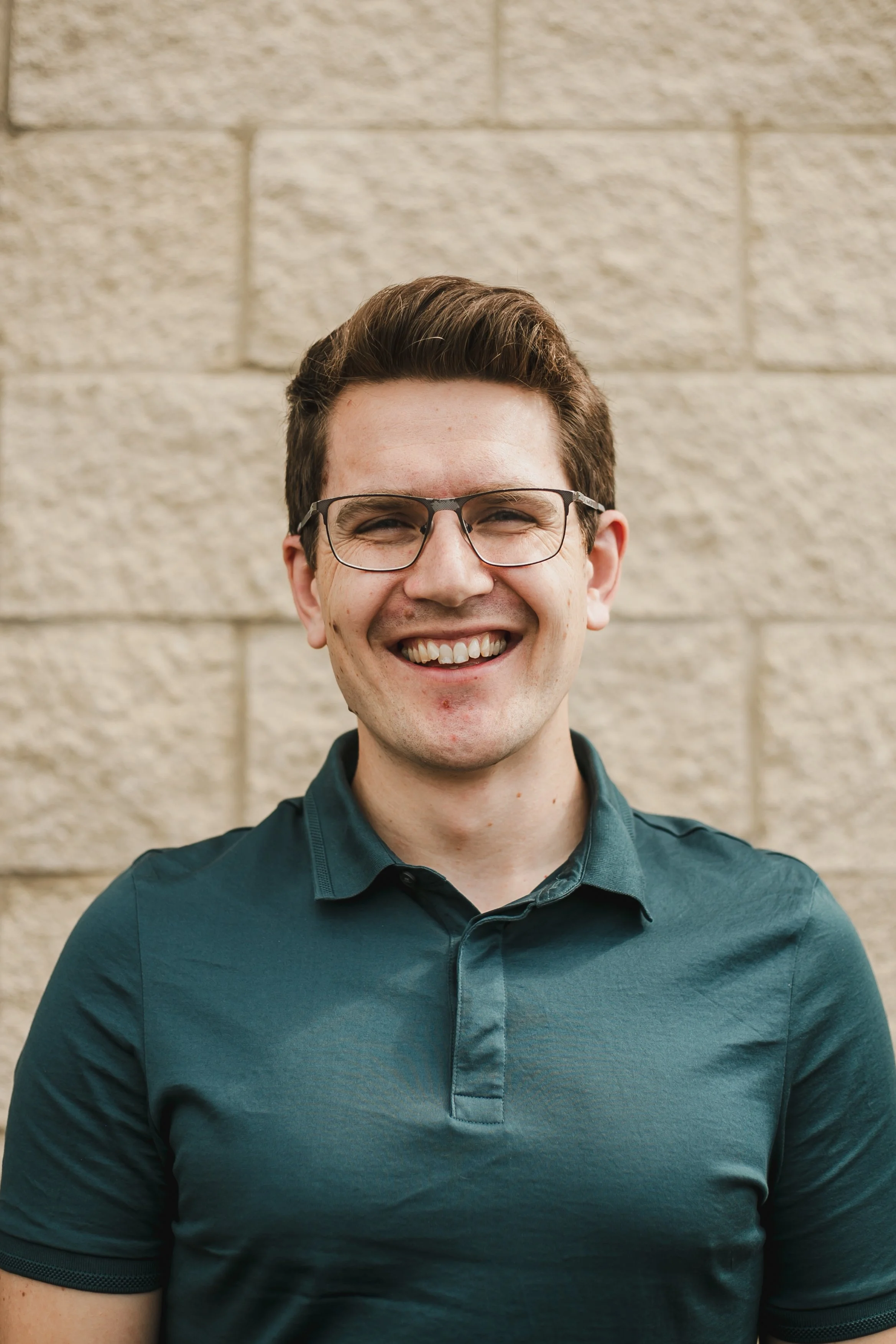Laurence Pallant. A young man smiling, wearing glasses and a teal collared shirt, standing in front of a beige brick wall.
