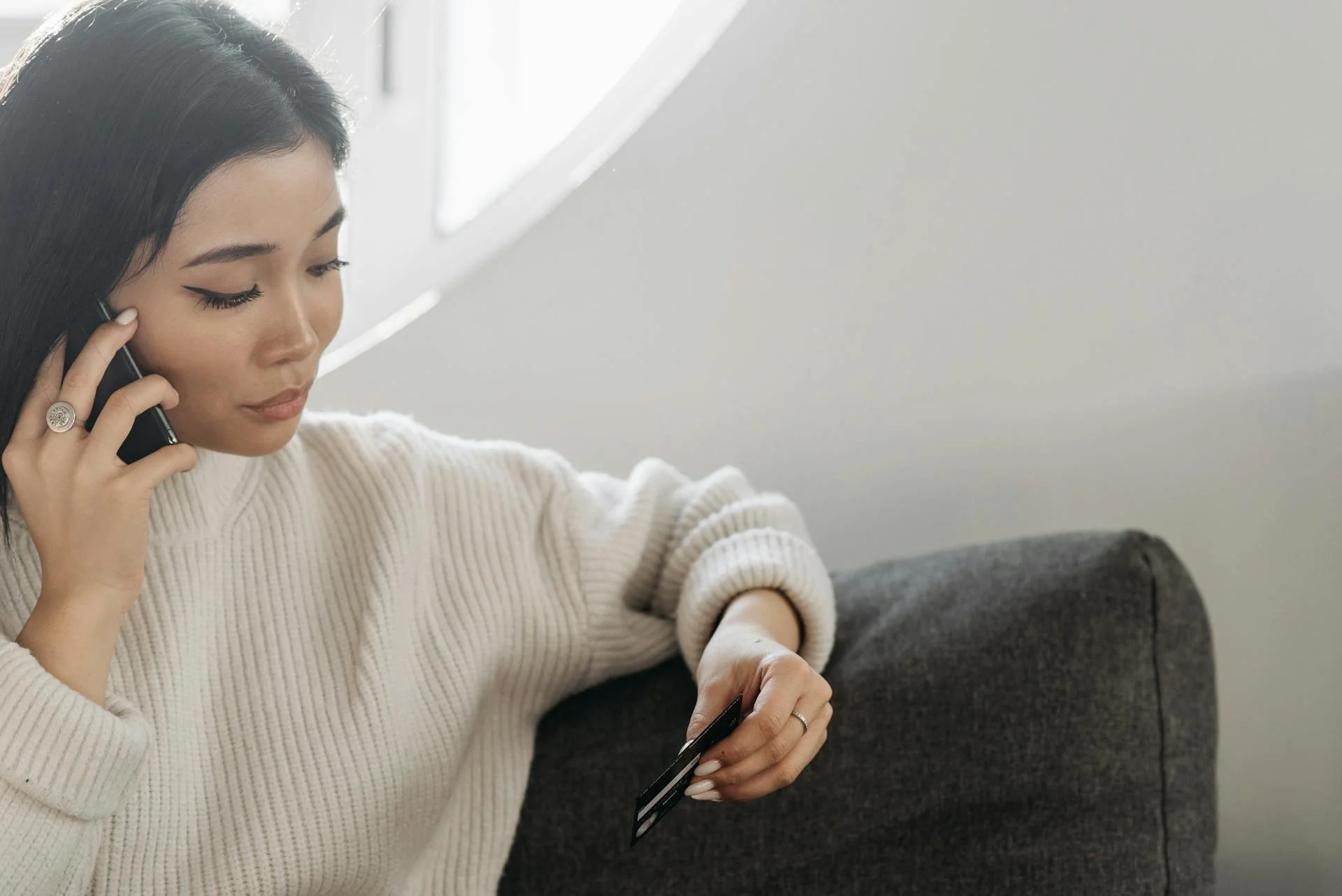 A woman with long dark hair, wearing a cream-colored sweater, sitting on a gray couch, talking on a mobile phone and holding a credit card.