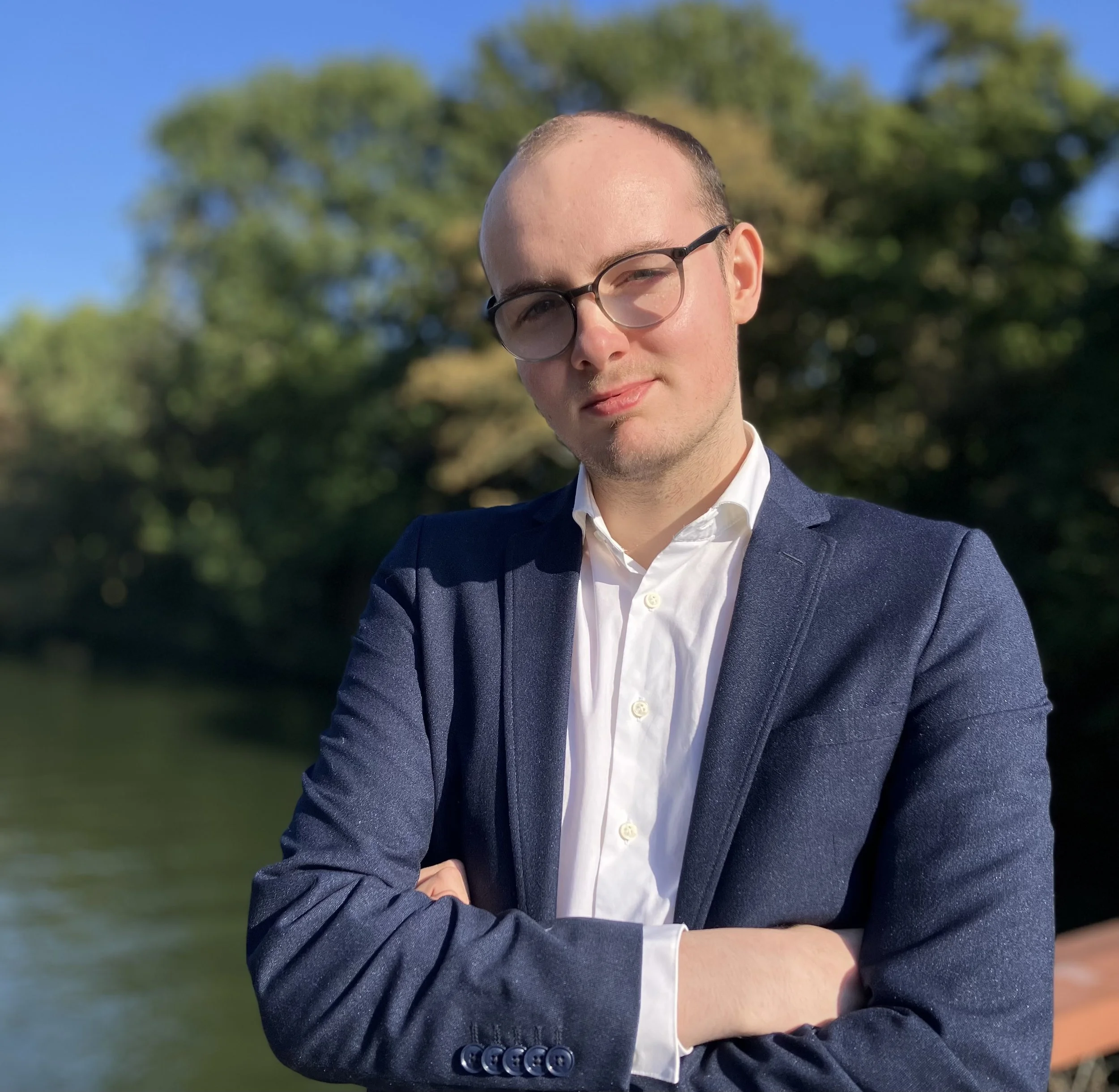 Portret van een jonge man met een kalende haardos, draagt een zwarte bril en een blauwe blazer, staand buiten met een groene boom en een blauwe lucht op de achtergrond.