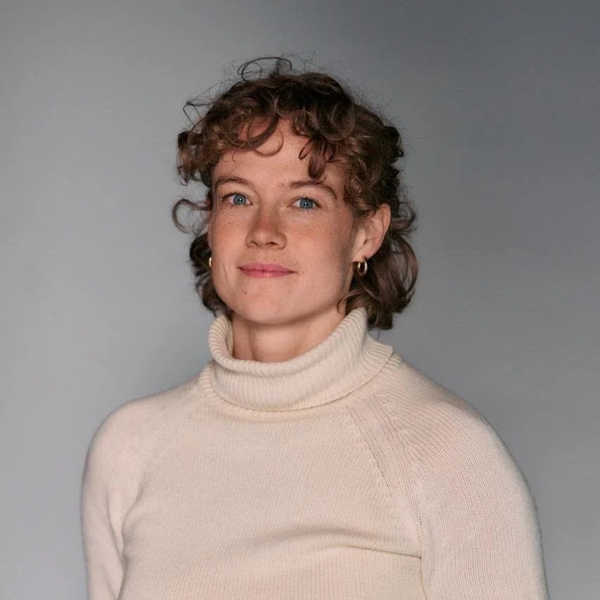 Portret van een vrouw met korte, gekrulde bruin haar, blauwe ogen, lichte huidskleur, die een witte coltrui draagt en een glimlach heeft, tegen een grijze achtergrond.