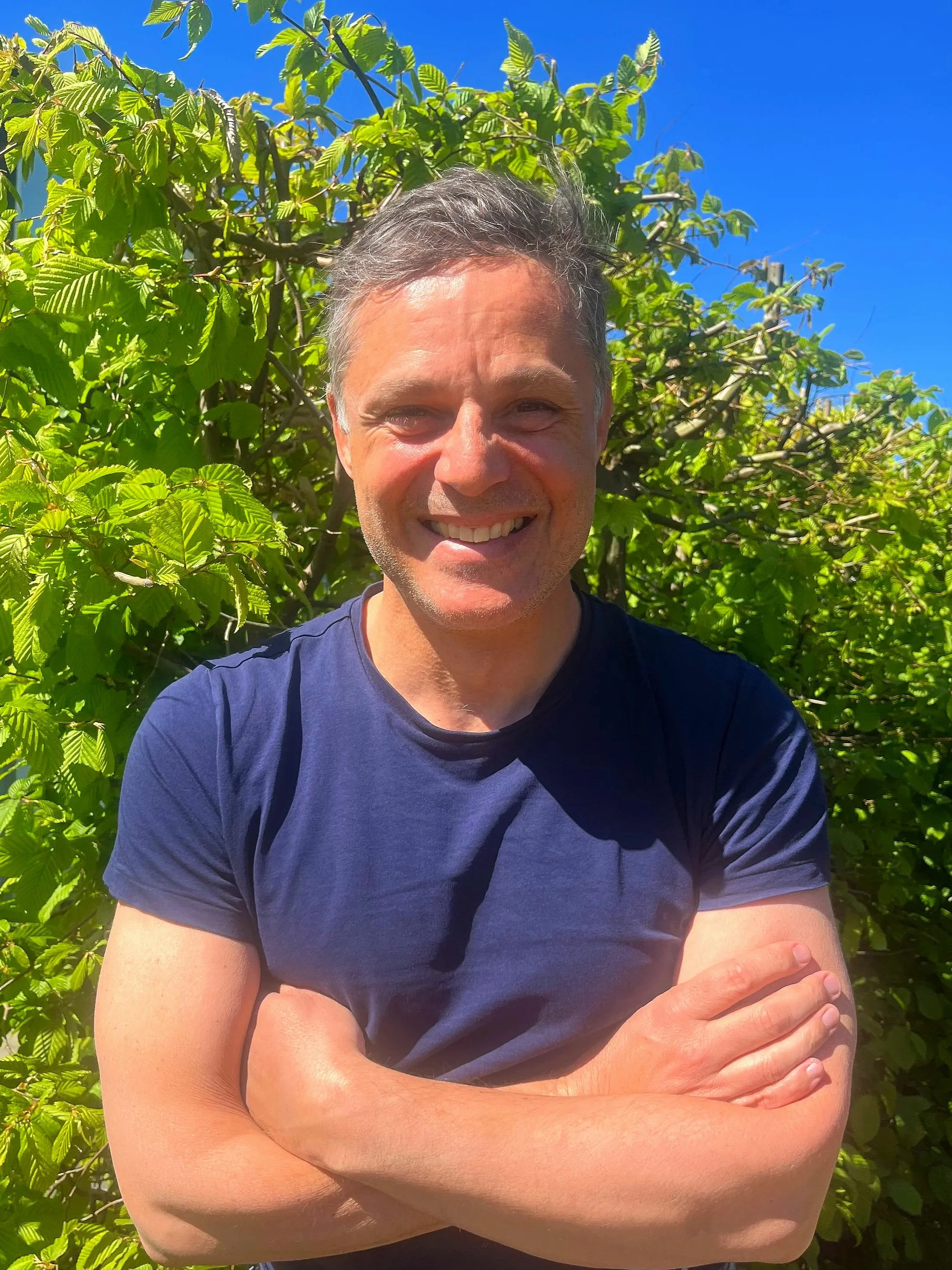 A man smiling with arms crossed outdoors, standing in front of green foliage and a blue sky.