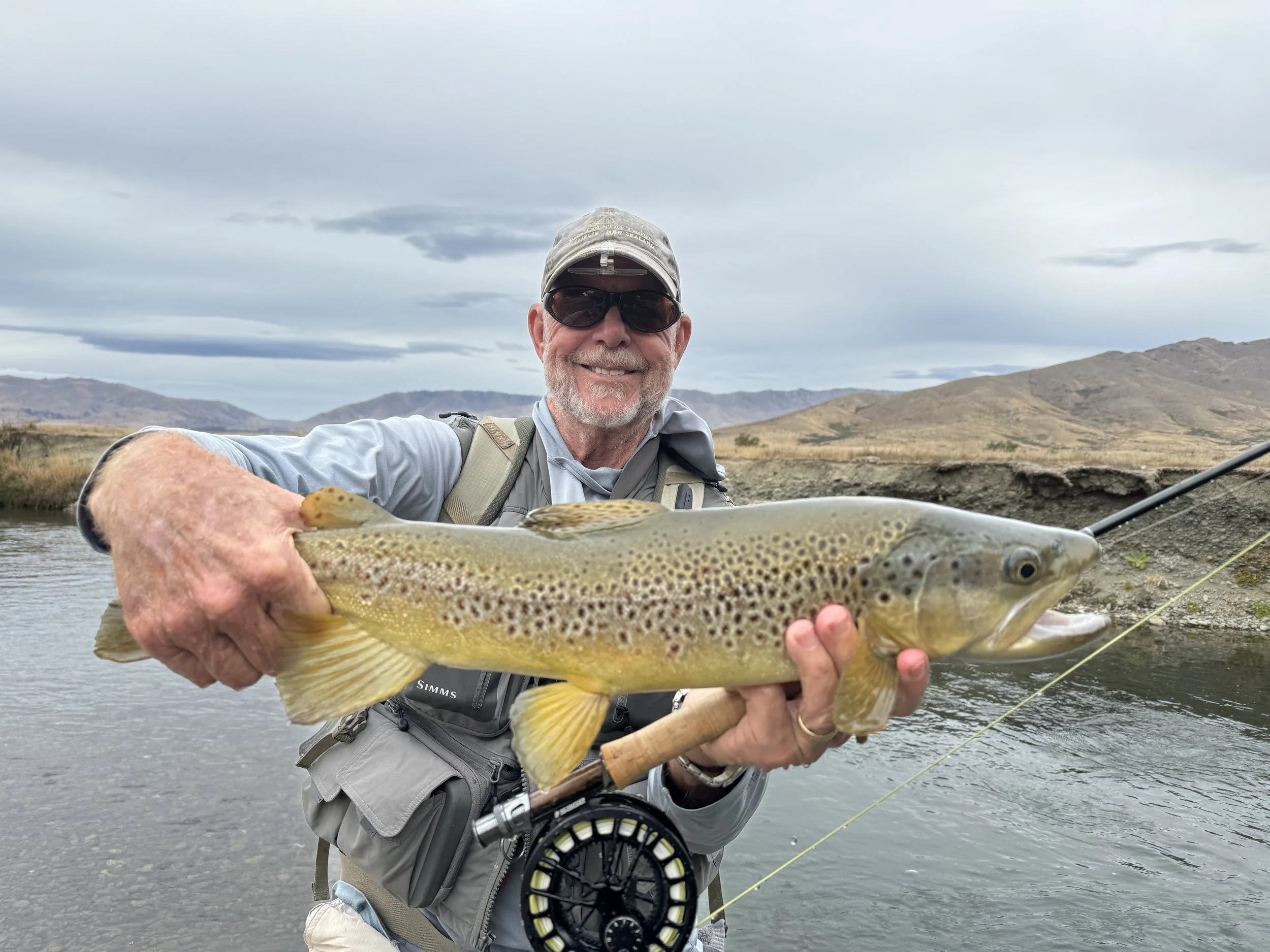 Man holding a large rainbow trout he caught while fishing in a river on a cloudy day, with mountains in the background.