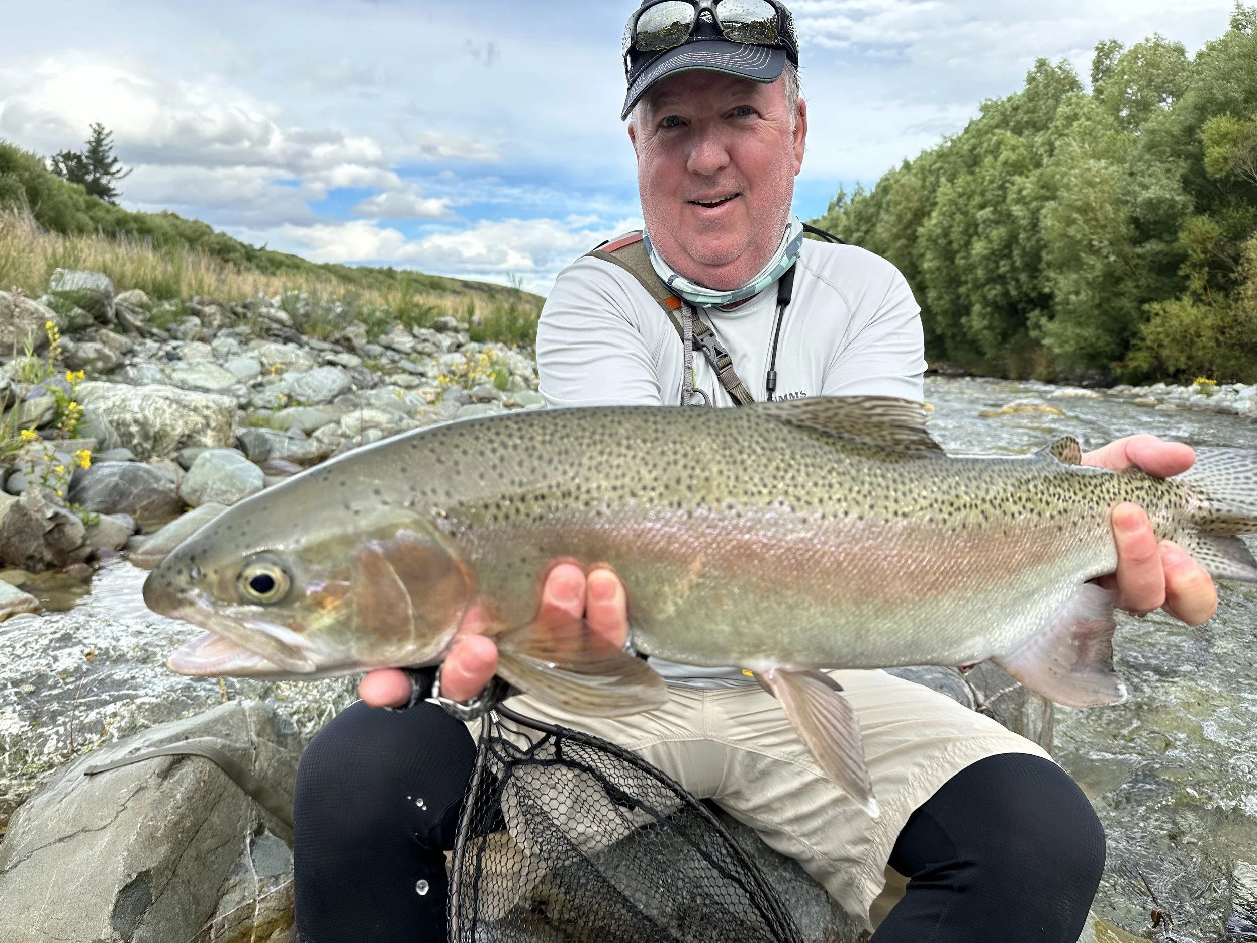 Man in outdoor gear holding a large rainbow trout fish over a rocky stream, with trees and cloudy sky in the background.