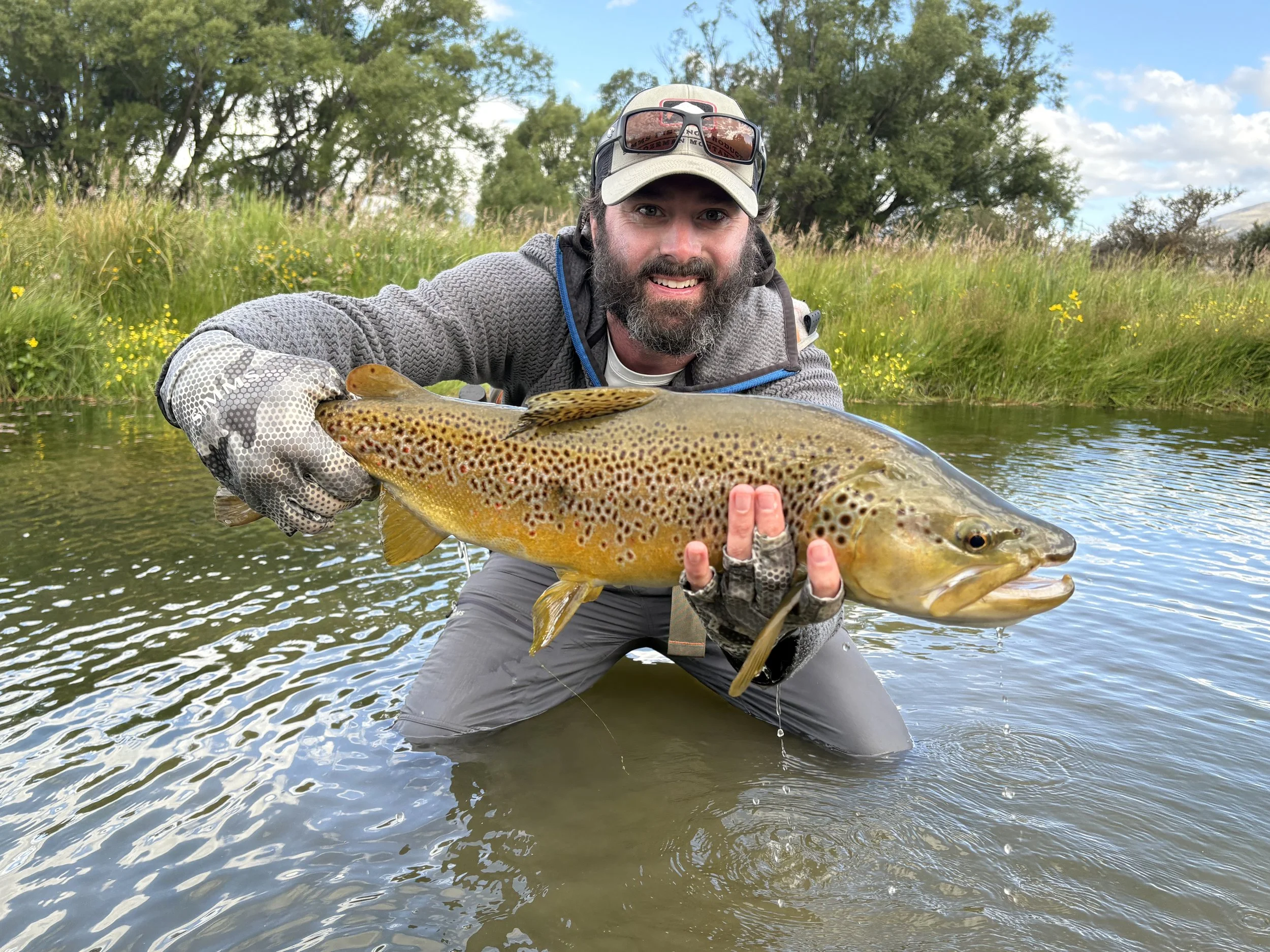 Man wearing a cap, sunglasses on his head, and fishing gloves, kneeling in a river, holding a large spotted trout with both hands, smiling at the camera, with green trees and yellow flowers on the riverbank in the background.