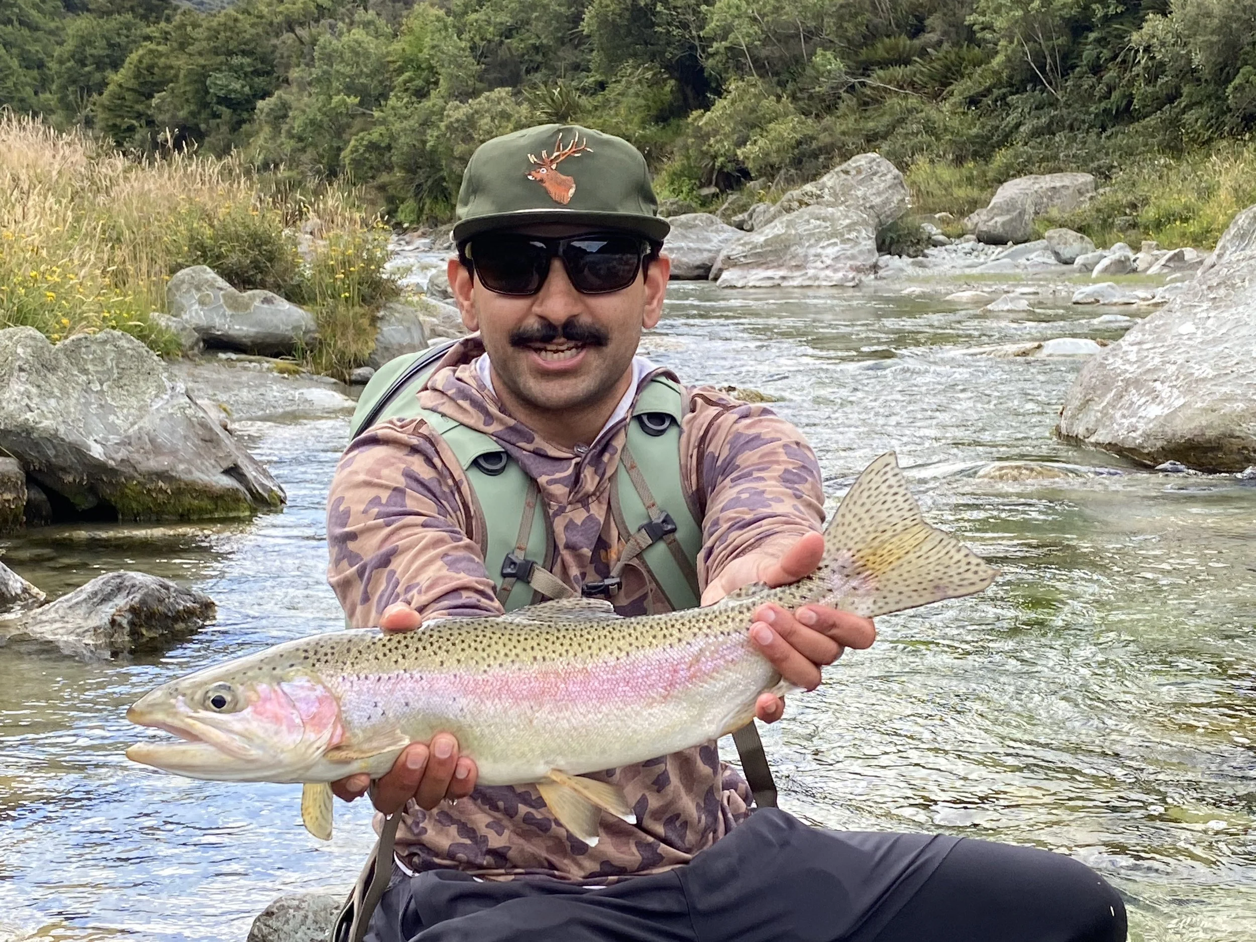 A man wearing sunglasses, a hat with a deer logo, and a camouflage shirt, sitting in a river and holding a large rainbow trout fish with both hands during daytime.