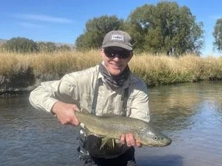 A person wearing sunglasses, a gray cap, and outdoor clothing holding a large fish by a river bank.