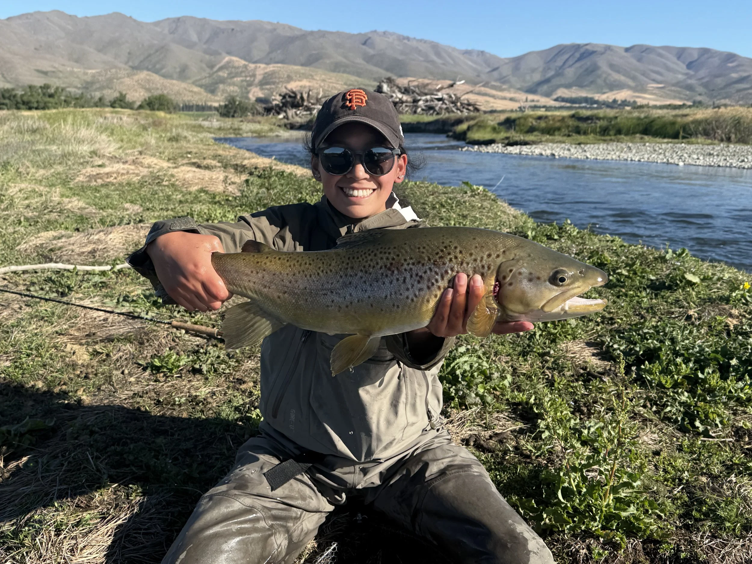 Person kneeling on grass by river holding a large brown trout with a smile, wearing sunglasses, a cap, and outdoor clothing, with mountains in the background.