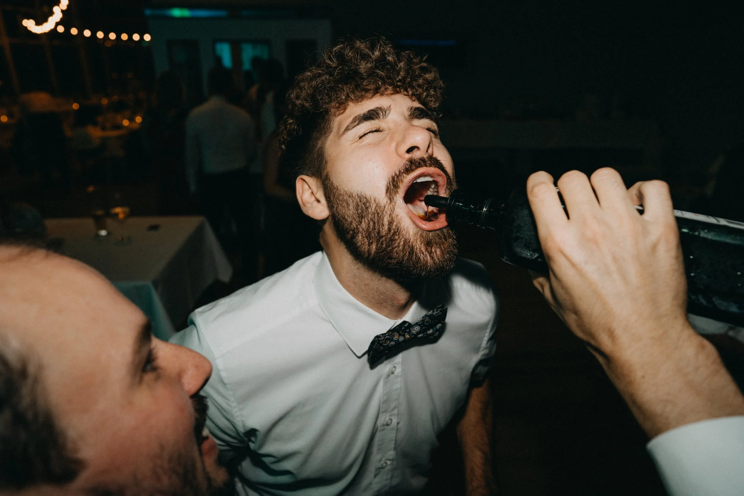 Ein Mann mit dunklen, lockigen Haaren und Bart trägt einen weißen Hemd und eine Fliege, während er singt oder spricht, umgeben von anderen Personen bei einer Feier in einer dunklen Umgebung.