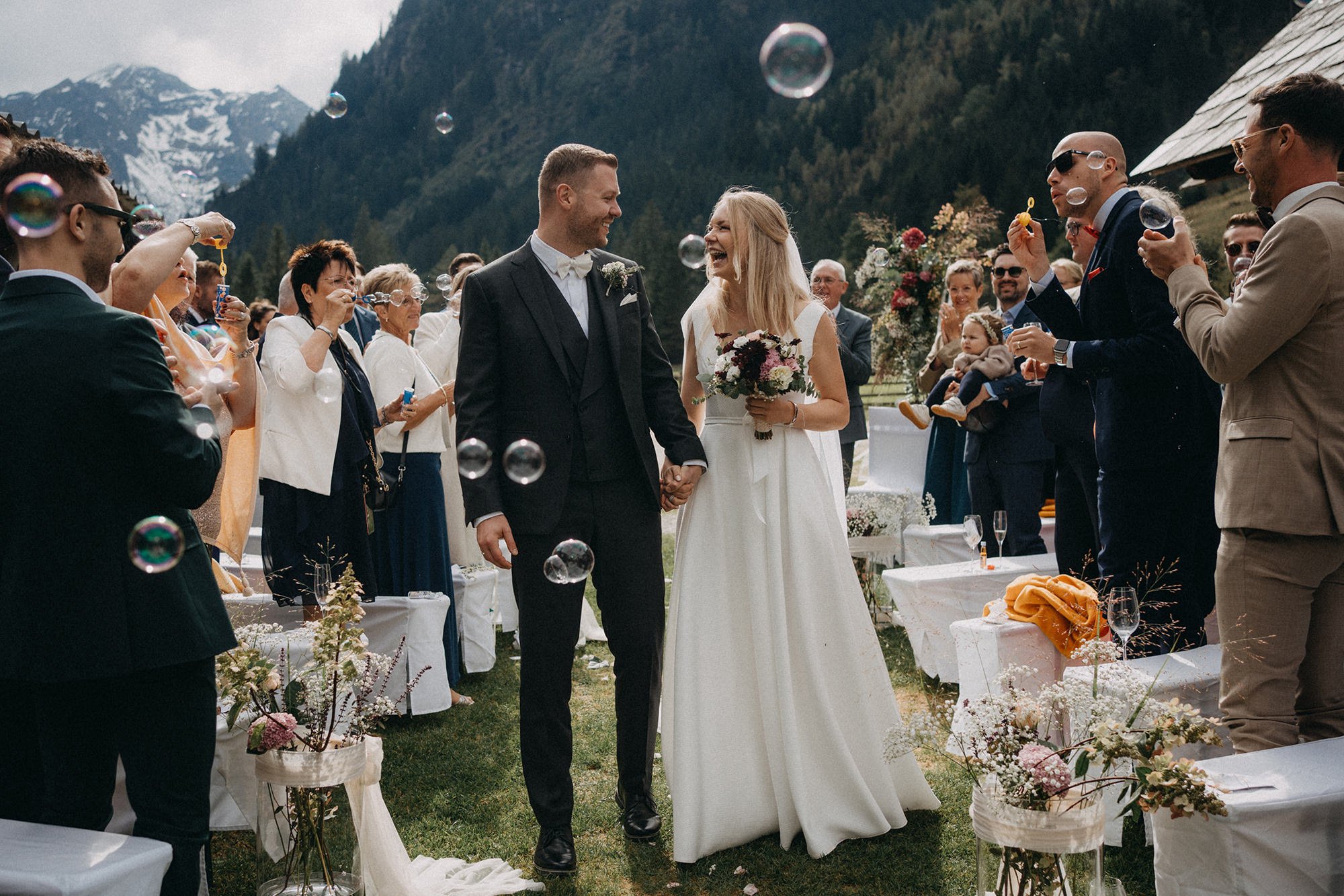 Ein Heiratsfest im Freien inmitten der Berge, bei dem ein Brautpaar sich anlächelt, umgeben von Gästen, die Seifenblasen machen, und festlich gedeckten Tischen mit Blumen und Dekorationen.