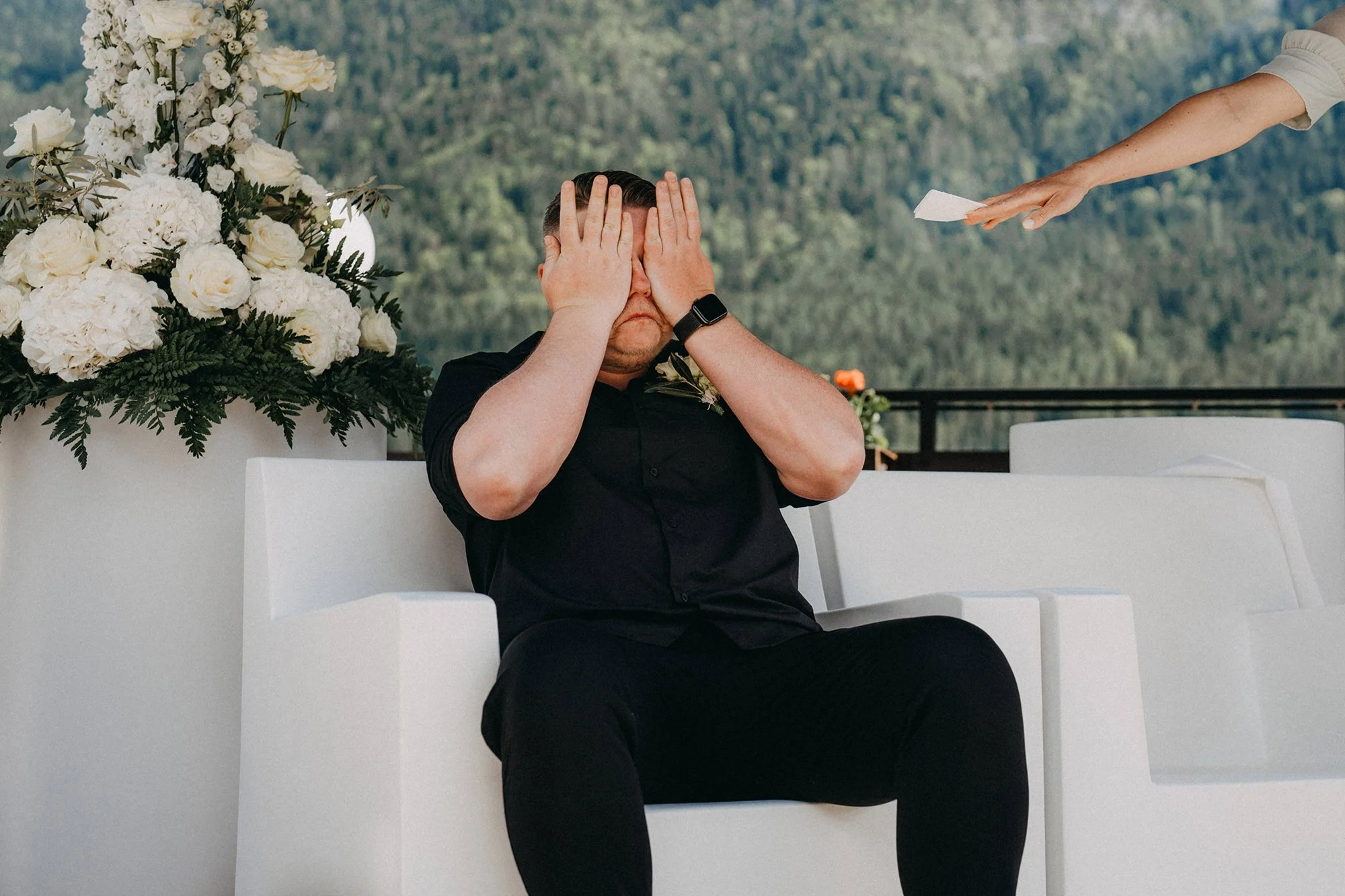 Ein Mann sitzt auf einer weißen Bank vor einem Blumenarrangement aus weißen Blumen und grünen Zweigen, während eine Person rechts den Arm ausstreckt, um an der Stelle, an der der Mann seine Hände an seinem Gesicht hat, eine rote Linie oder Träne zu zeichnen.