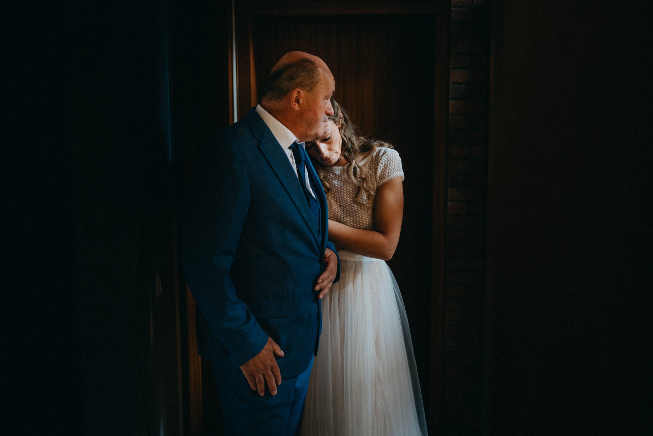 Ein Mann in einem blauen Anzug und eine Frau in einem weißen Kleid stehen eng nebeneinander in einem dunklen Raum, scheinen einen Moment der Trauer oder des Trostes zu teilen.