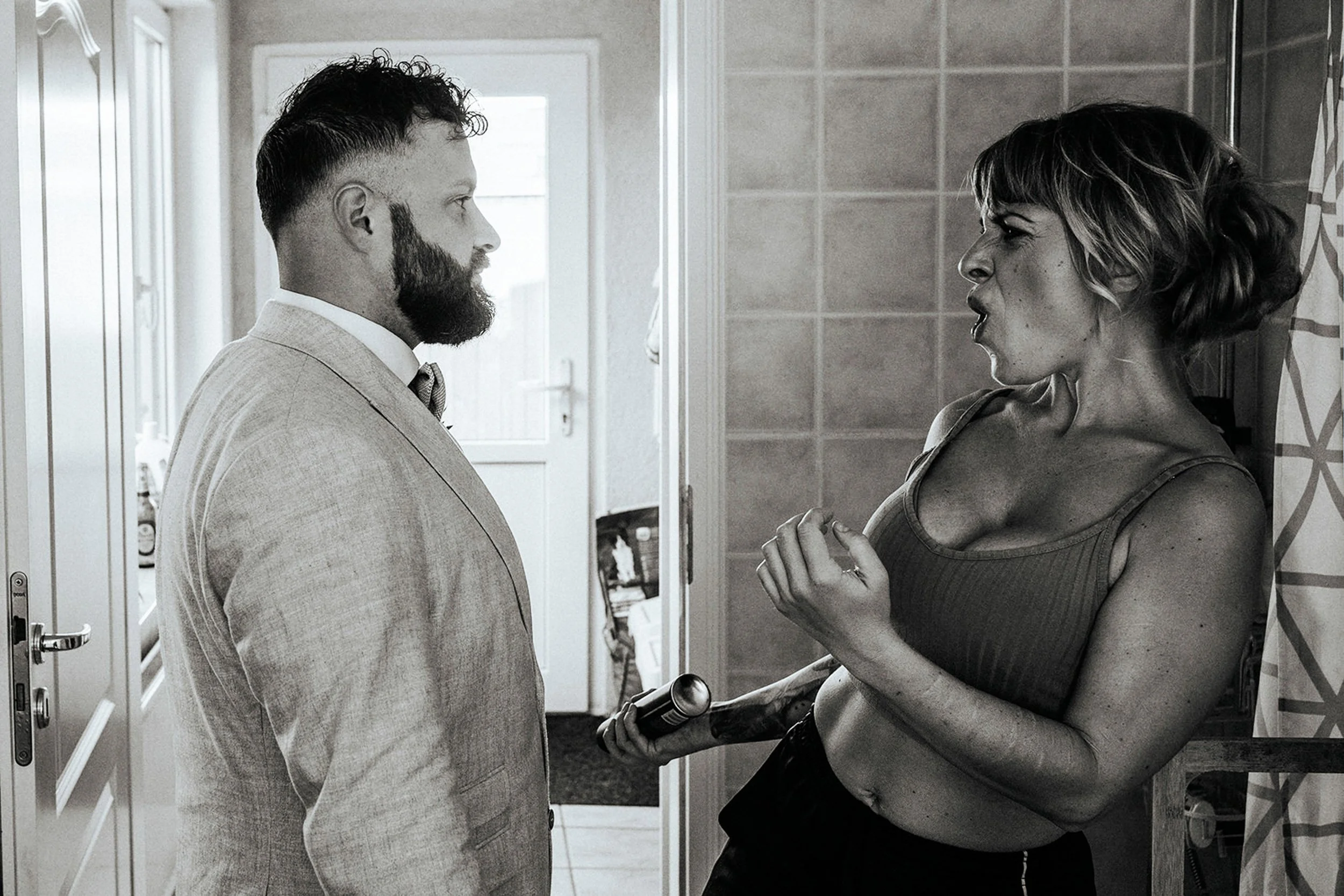 Eine schwarze und weiße Aufnahme eines Streits zwischen einem Mann und einer Frau in einer begehbaren Dusche. Der Mann trägt einen Anzug und hat einen Bart, während die Frau eine ärmellose Bluse trägt und gestikulierend ist.