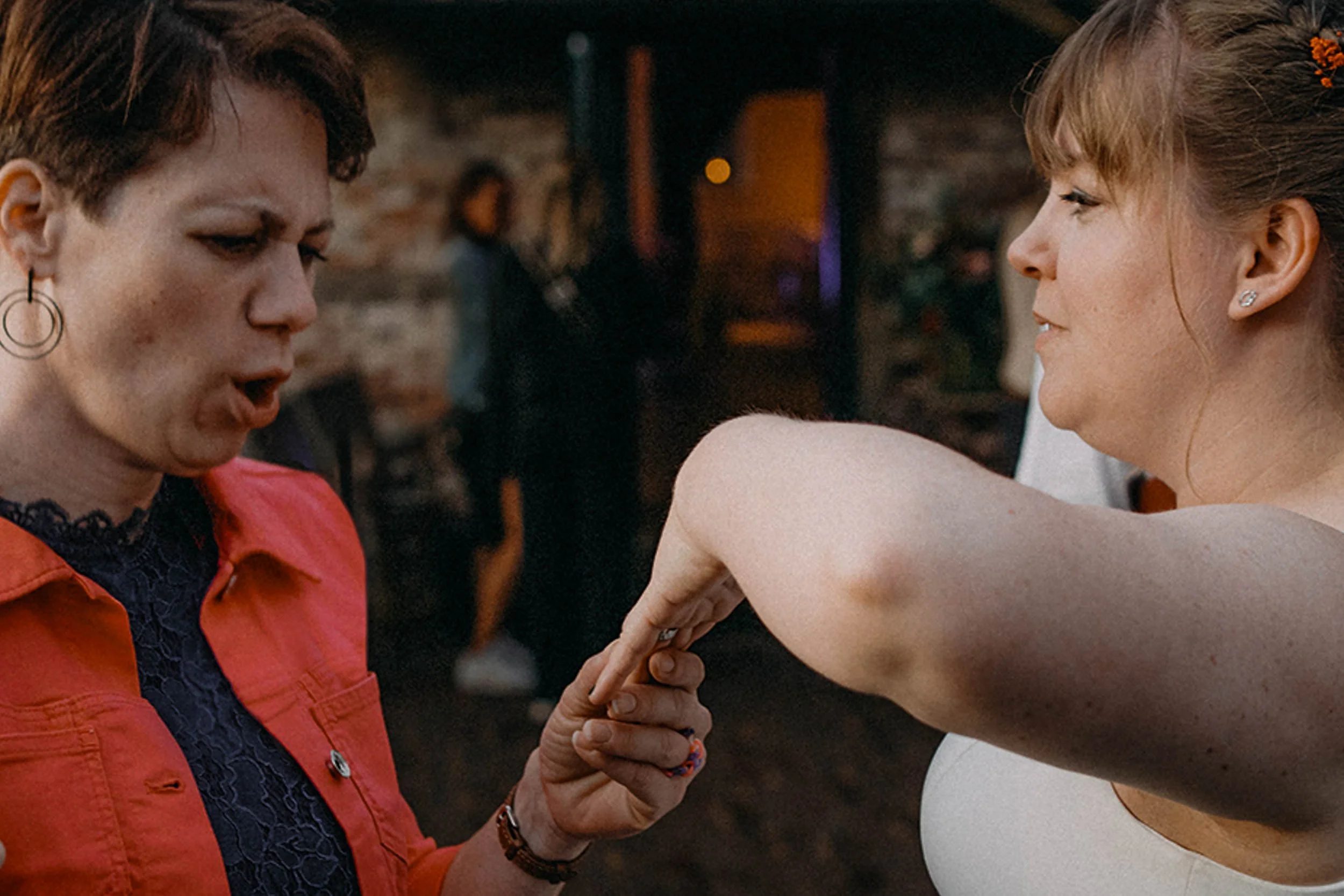 Zwei Frauen bei einer Armwrestling-Kampf, eine trägt ein rotes Hemd, die andere einen weißen Top, im Hintergrund eine Partyszene.