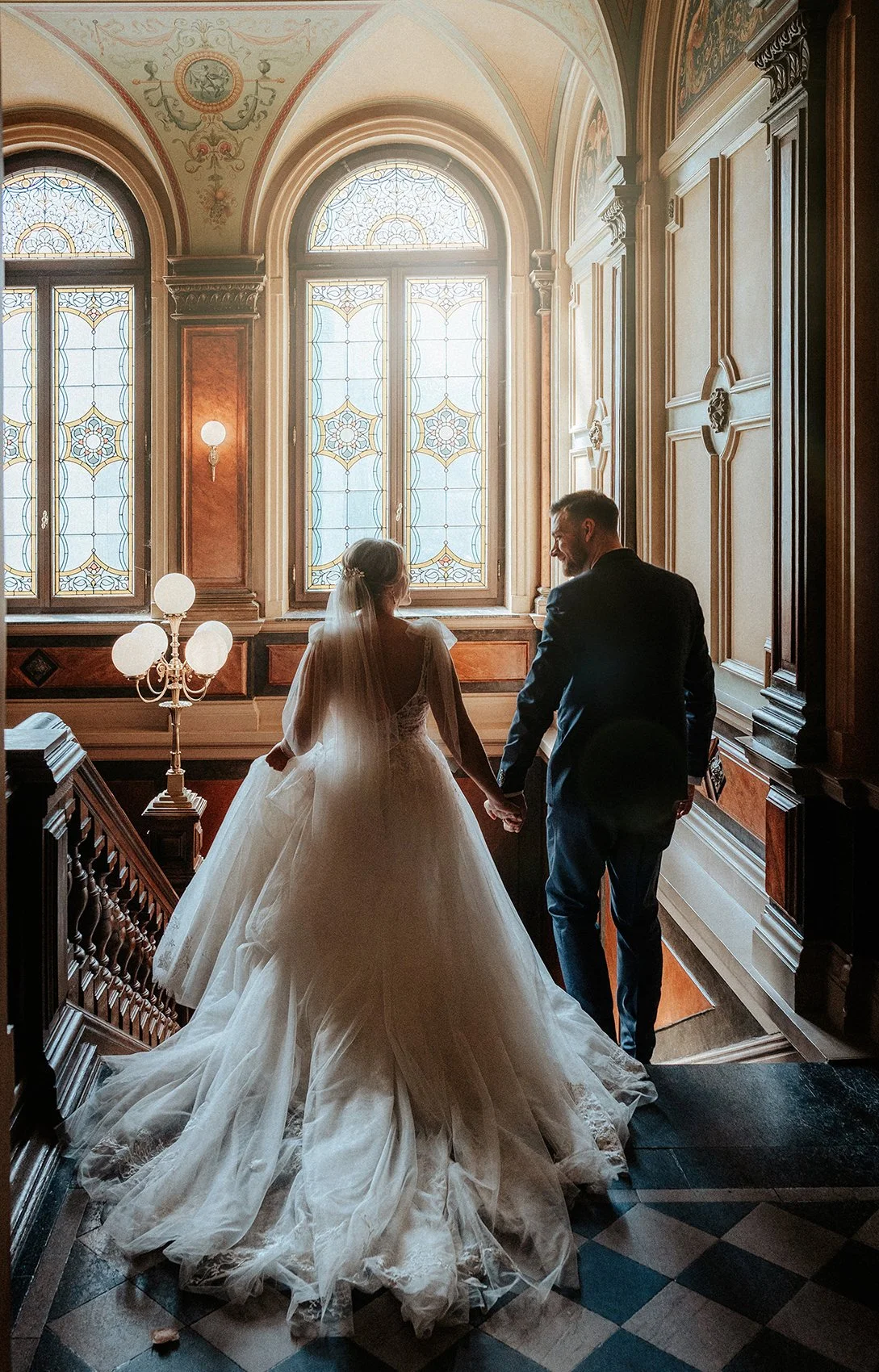 Ein Brautpaar in einem eleganten Saal mit großen bunten Fenstern, die viel Licht hereinlassen. Die Braut trägt ein voluminöses weißes Hochzeitskleid, der Bräutigam einen dunklen Anzug. Sie halten sich an den Händen, während sie die Treppe hinauflaufen.