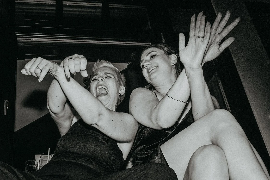 Zwei Frauen in einem schwarzen Kleid, die lachen und sich umarmen, in einer dunklen Umgebung.