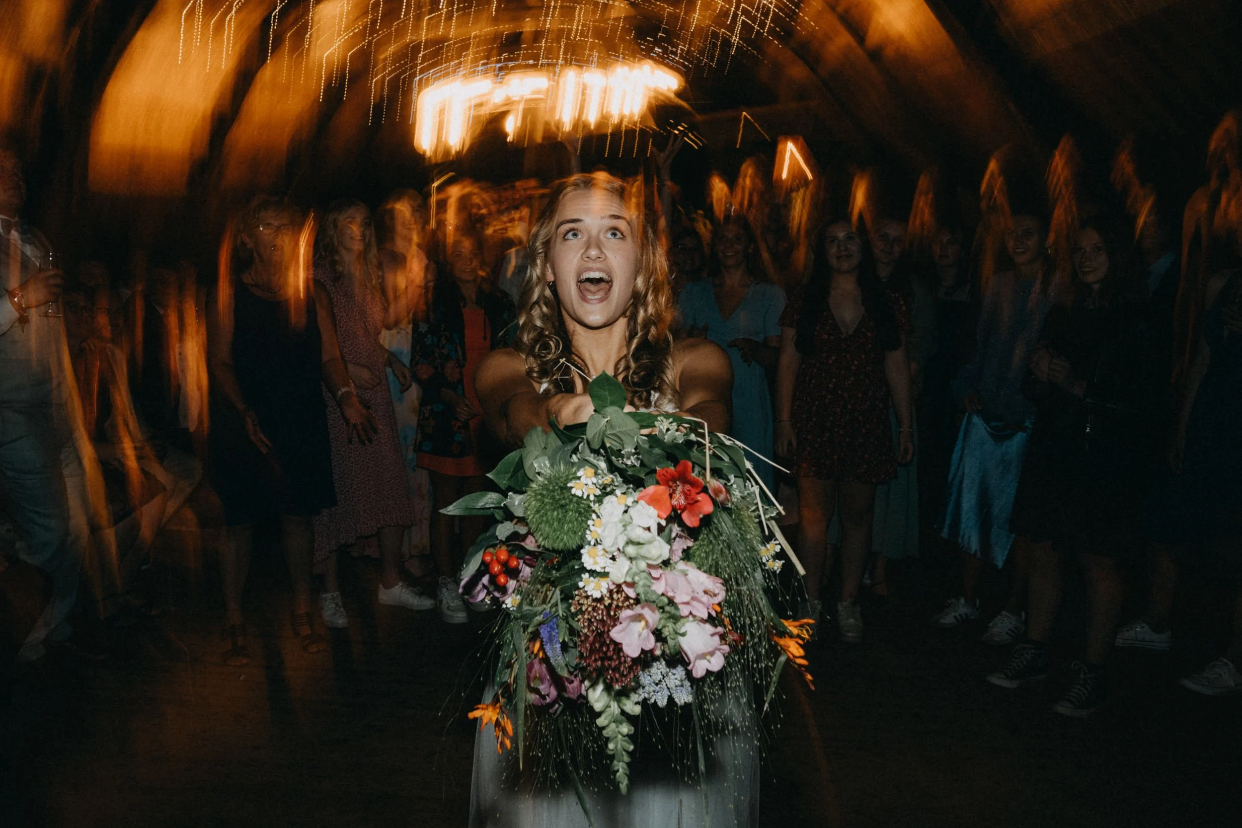 Eine junge Frau mit einem Blumenstrauß auf einem Fest, umgeben von einer Gruppe von lachenden Menschen, mit bewegter Beleuchtung im Hintergrund.