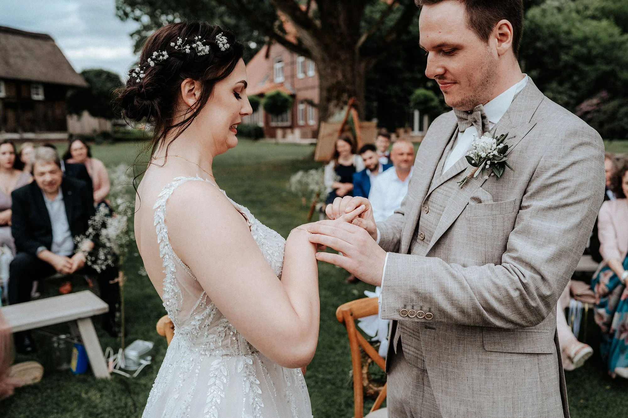 Braut und Bräutigam tauschen Trauringe bei ihrer Hochzeit im Garten, im Hintergrund sitzen Gäste und ein Baum im Freien.