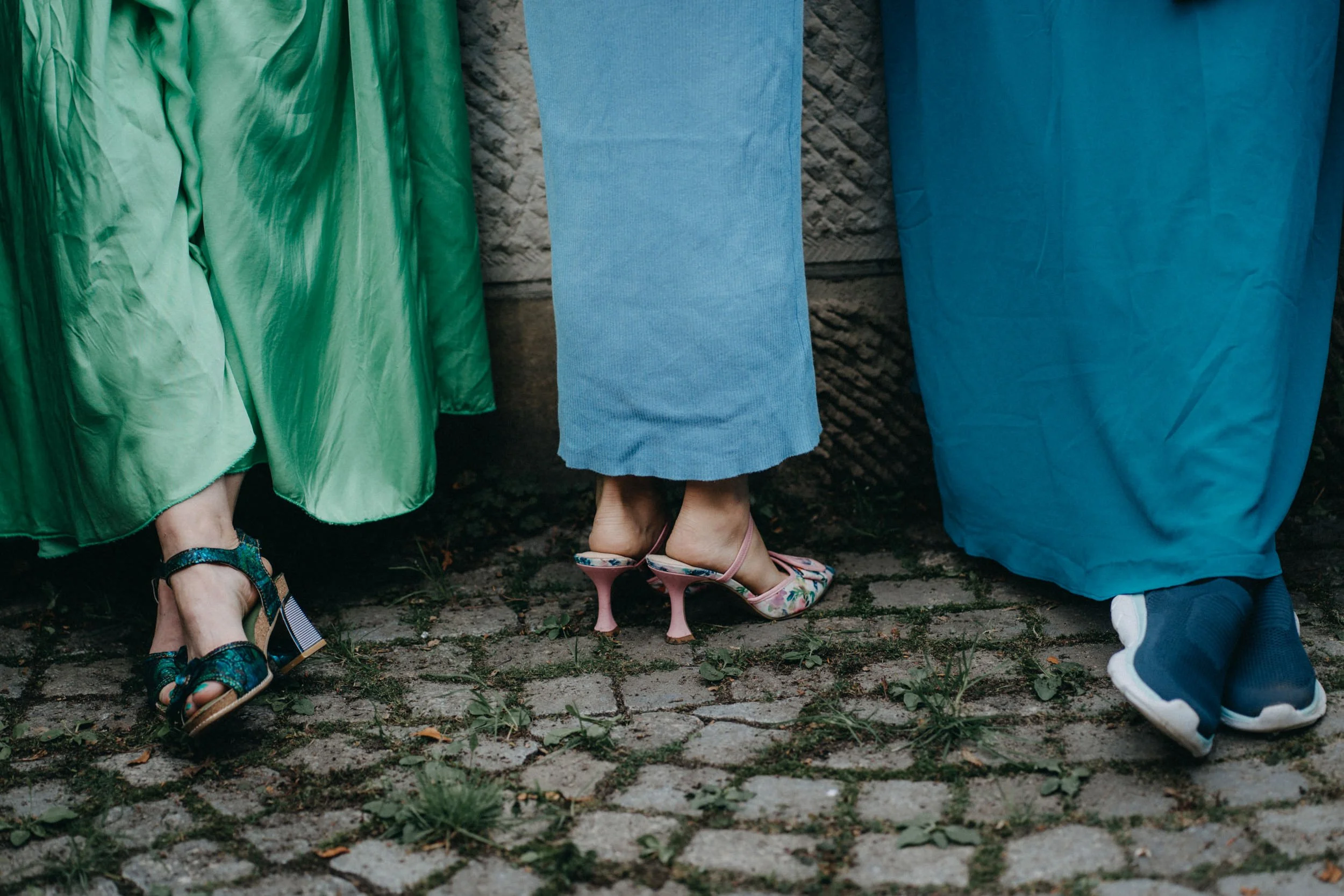Menschen stehen nebeneinander, nur die Beine sind sichtbar, sie tragen verschiedene Sandalen und Schuhe, auf einem gepflasterten Boden.