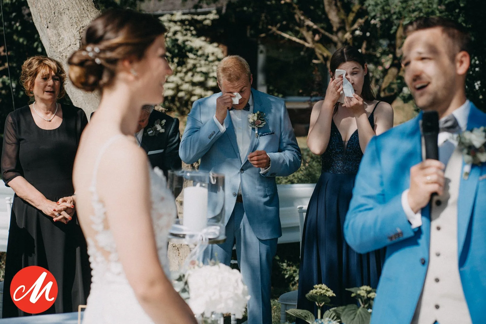 Trauung im Freien, Braut in Hochzeitskleid, Bräutigam in blauem Anzug, Familie und Freunde emotional bei der Zeremonie, eine Frau weint, eine andere hält Tuch vor dem Gesicht.
