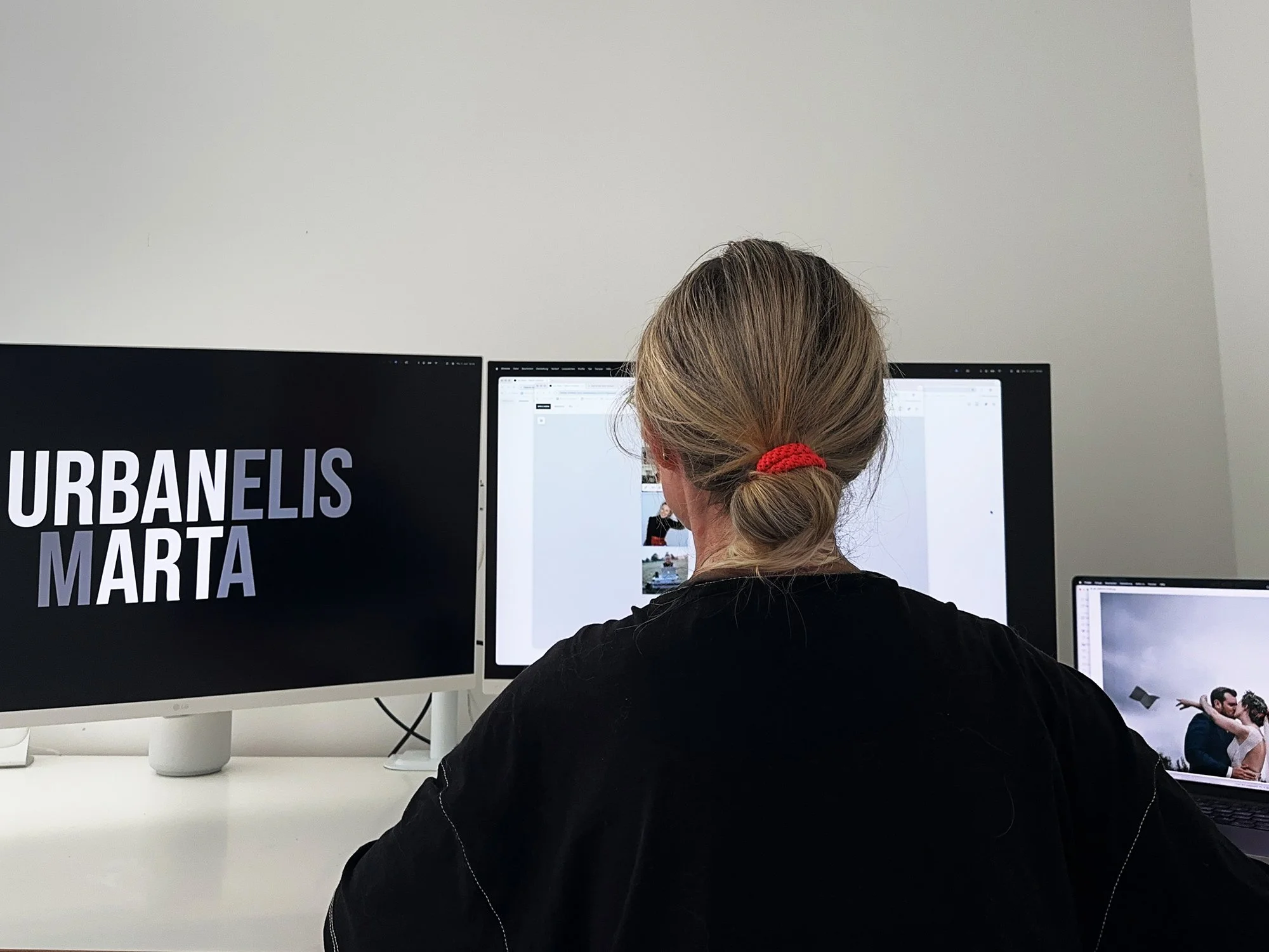 Person mit blondem Haar, gebunden mit rotem Haarband, sitzt vor mehreren Computerbildschirmen in einem modernen Büro, auf einem Bildschirm ist ein Text mit der Überschrift 'URBANELIS' sichtbar.