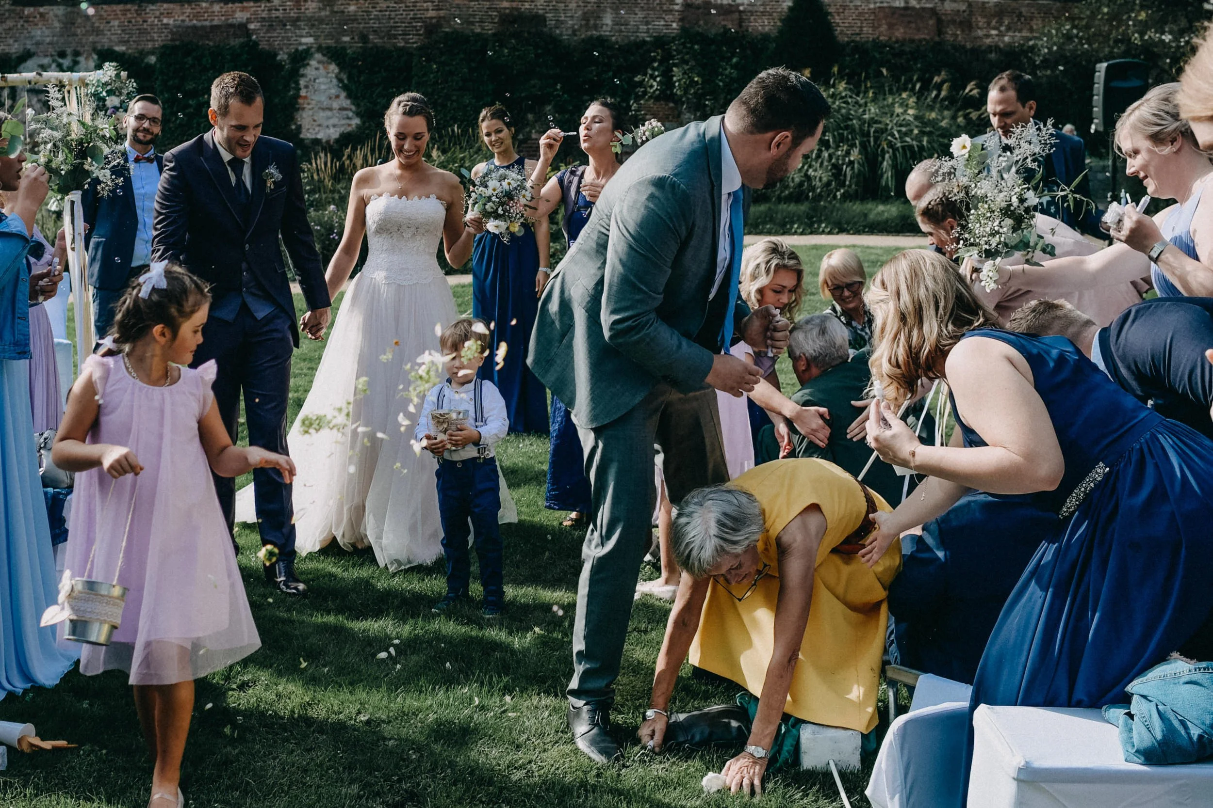 Hochzeitsparty im Garten, Gäste werfen Blüten, Braut und Bräutigam in der Mitte, umgeben von lachenden Freunden und Familie.