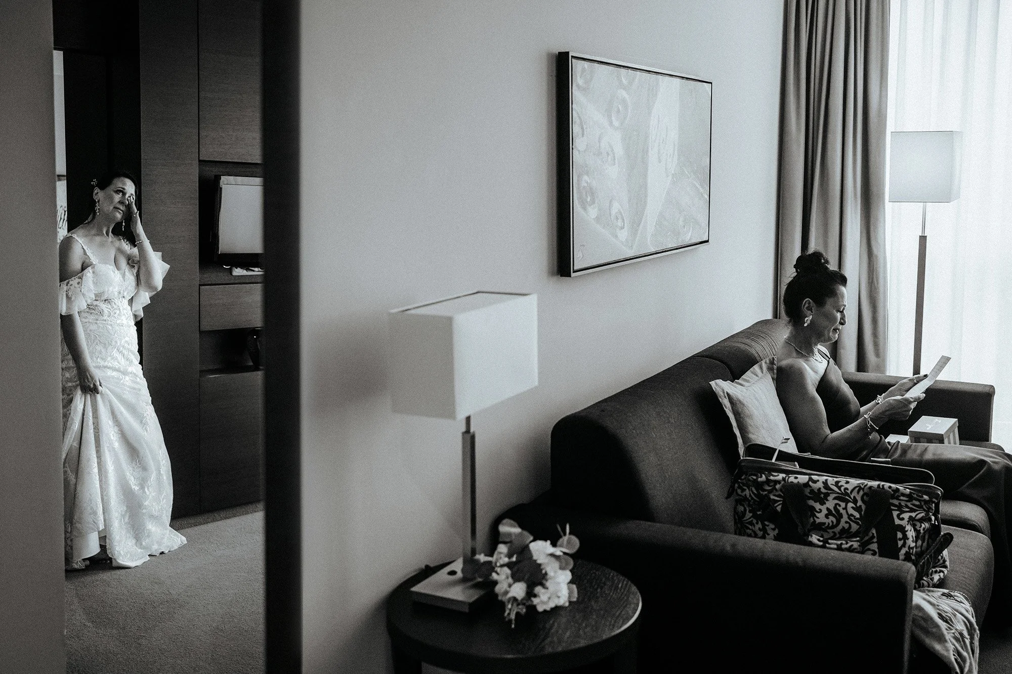 Zwei Frauen im Hotelzimmer: Eine Frau trägt ein brautkleid, die andere sitzt auf dem Sofa und liest eine Zeitschrift.