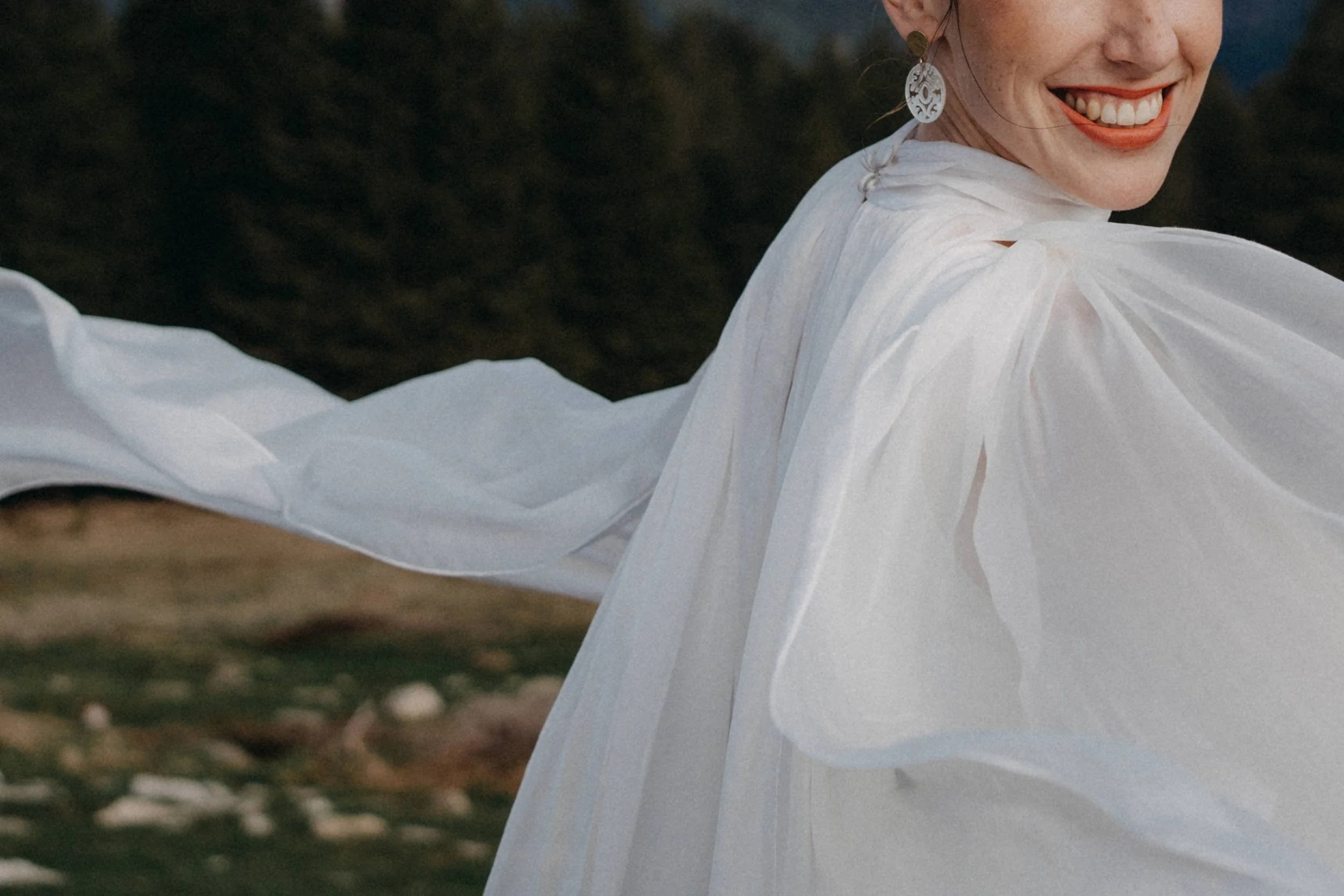 Nähe eines lachenden Damen mit roten Lippen, großen Ohrringen und weißem Kleid, im Freien bei Sonnenuntergang.