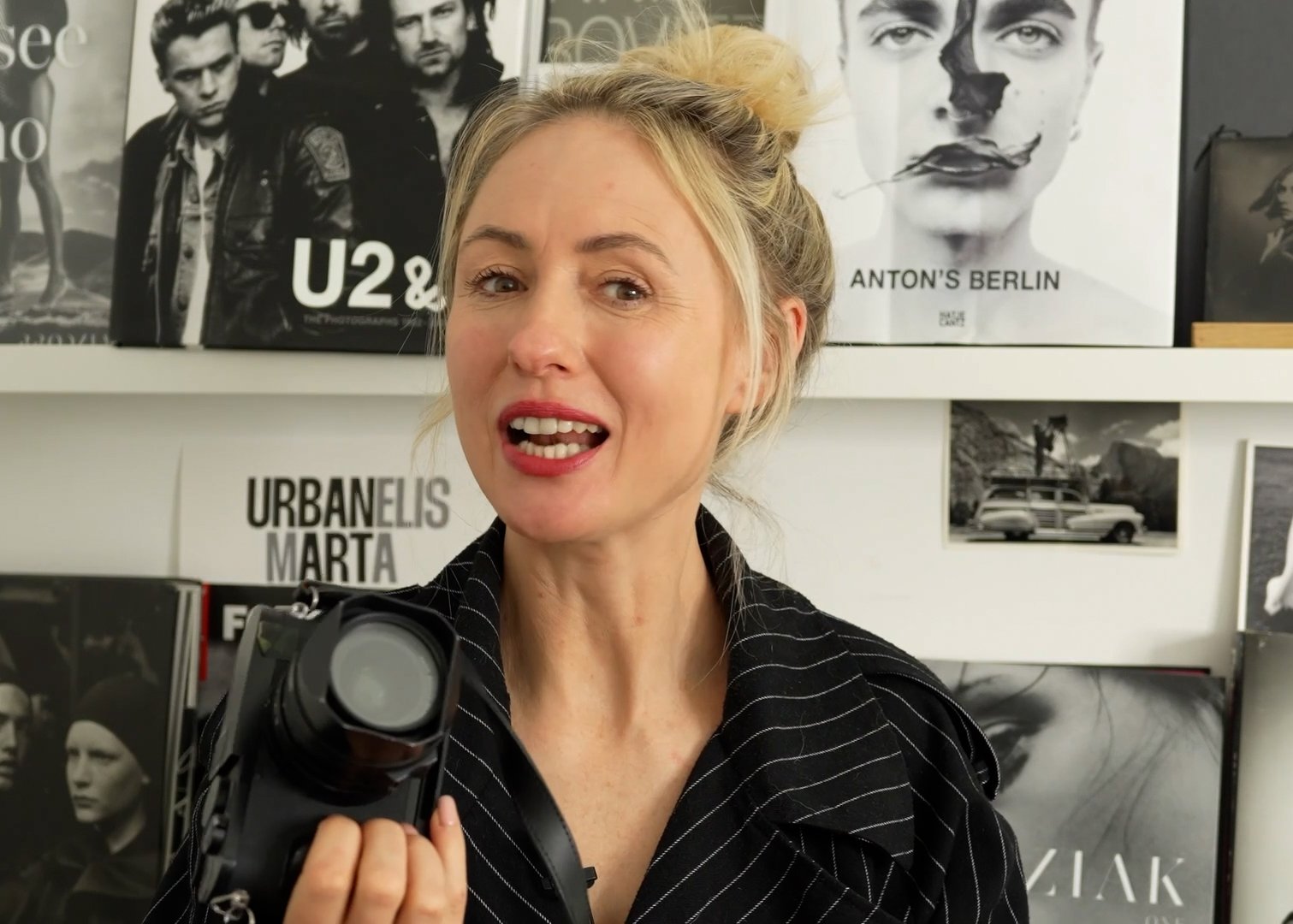 Eine blonde Frau hält eine Kamera in der Hand und schaut in die Kamera, im Hintergrund sind Plakate von Musikbands und eine schwarze und weiße Fotografie.